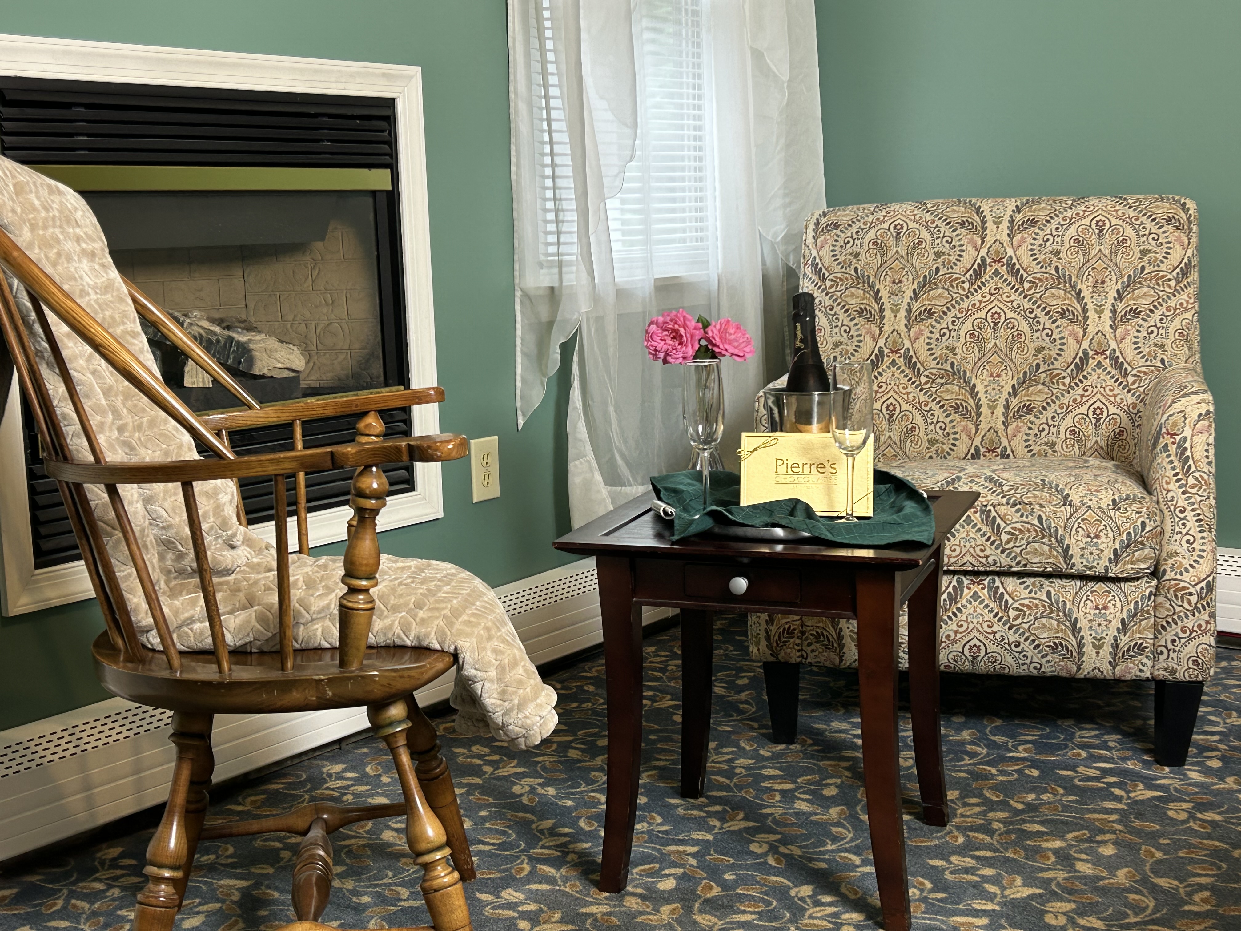 View of sitting area and ire place with 2 chairs, table, champagne, 2 glasses and box of chocolates
