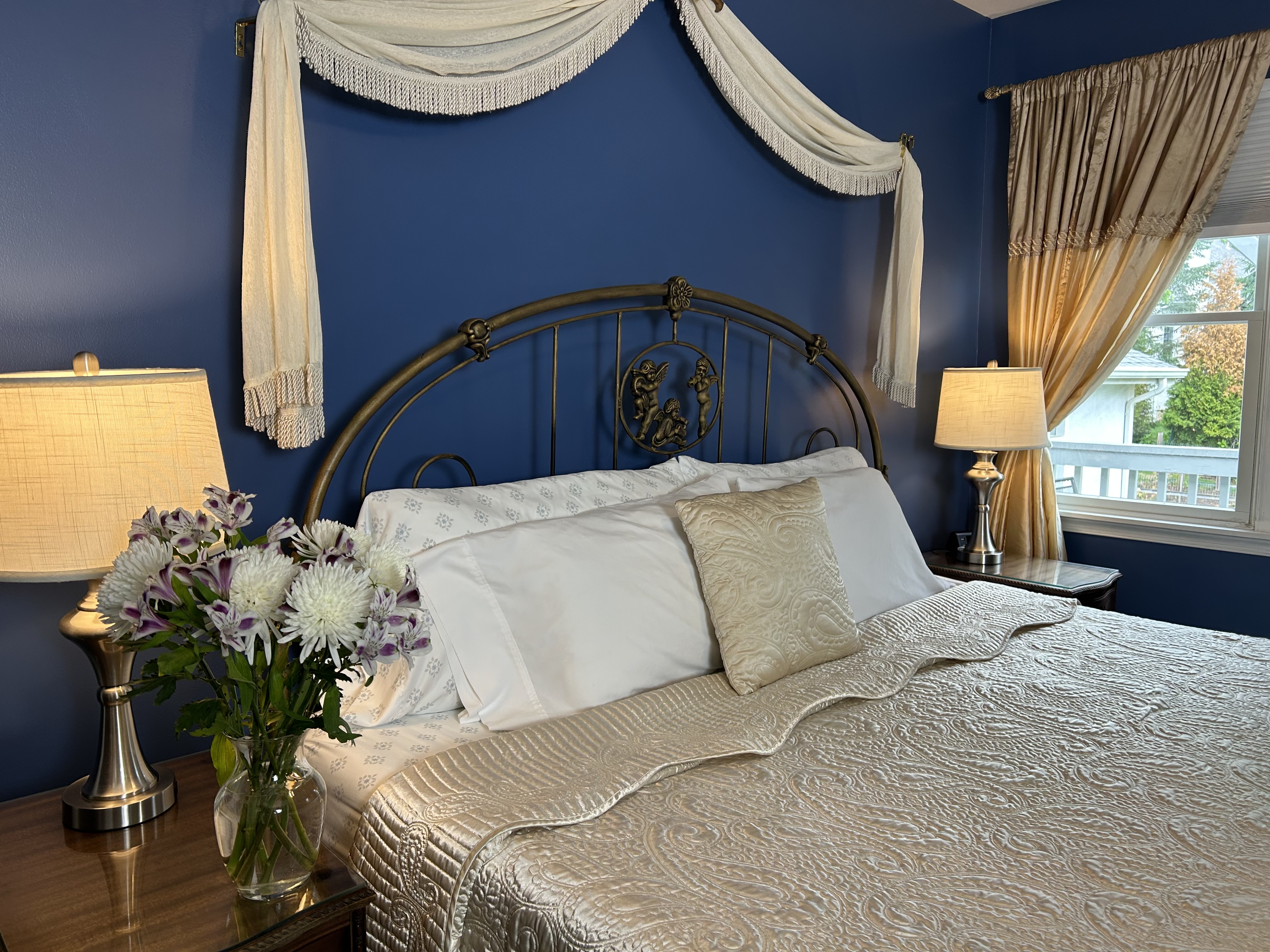 View of king bed with a side table, arm chair, flowers and a window