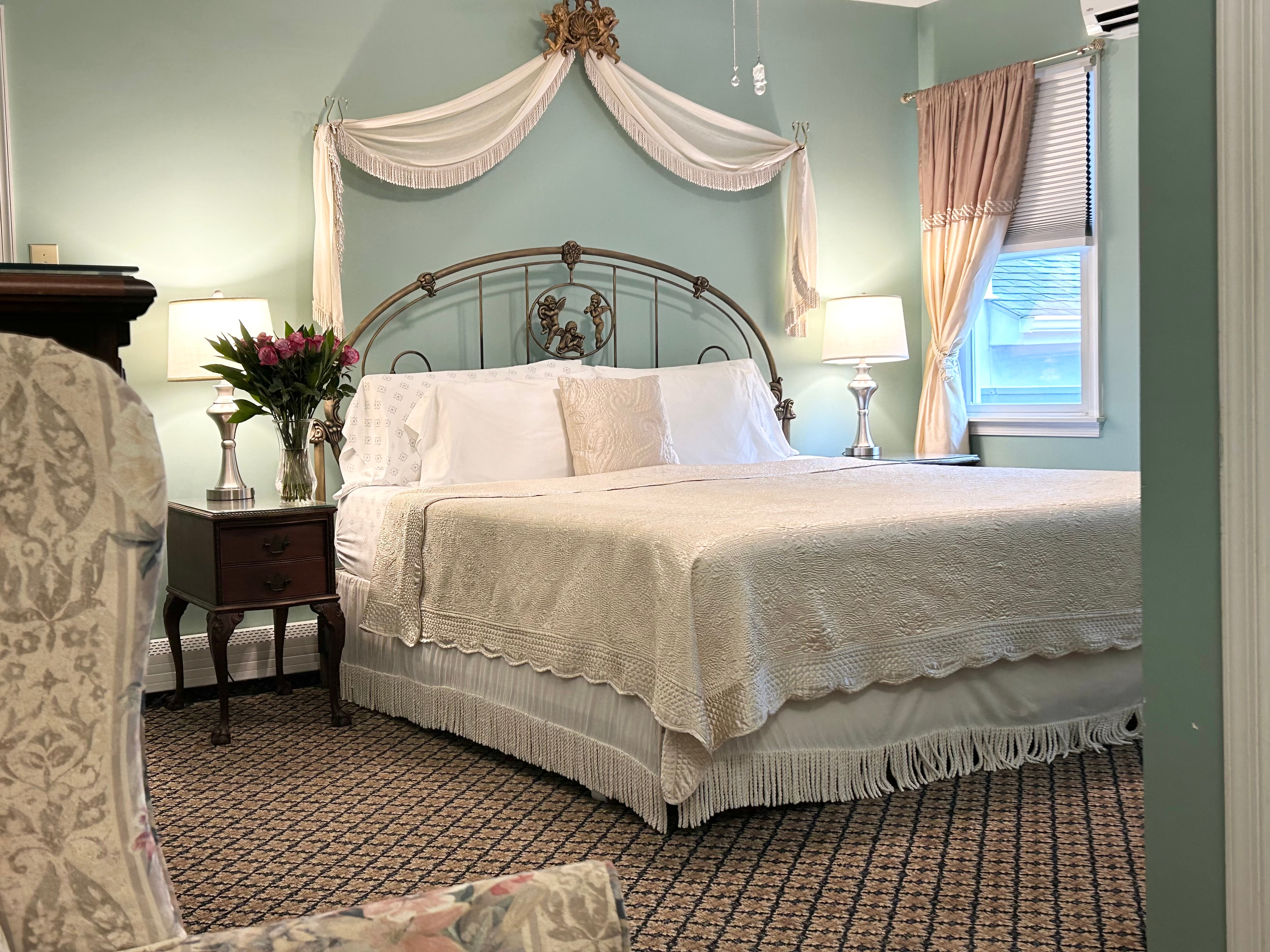 View of king bed with a side table, arm chair, flowers and a window