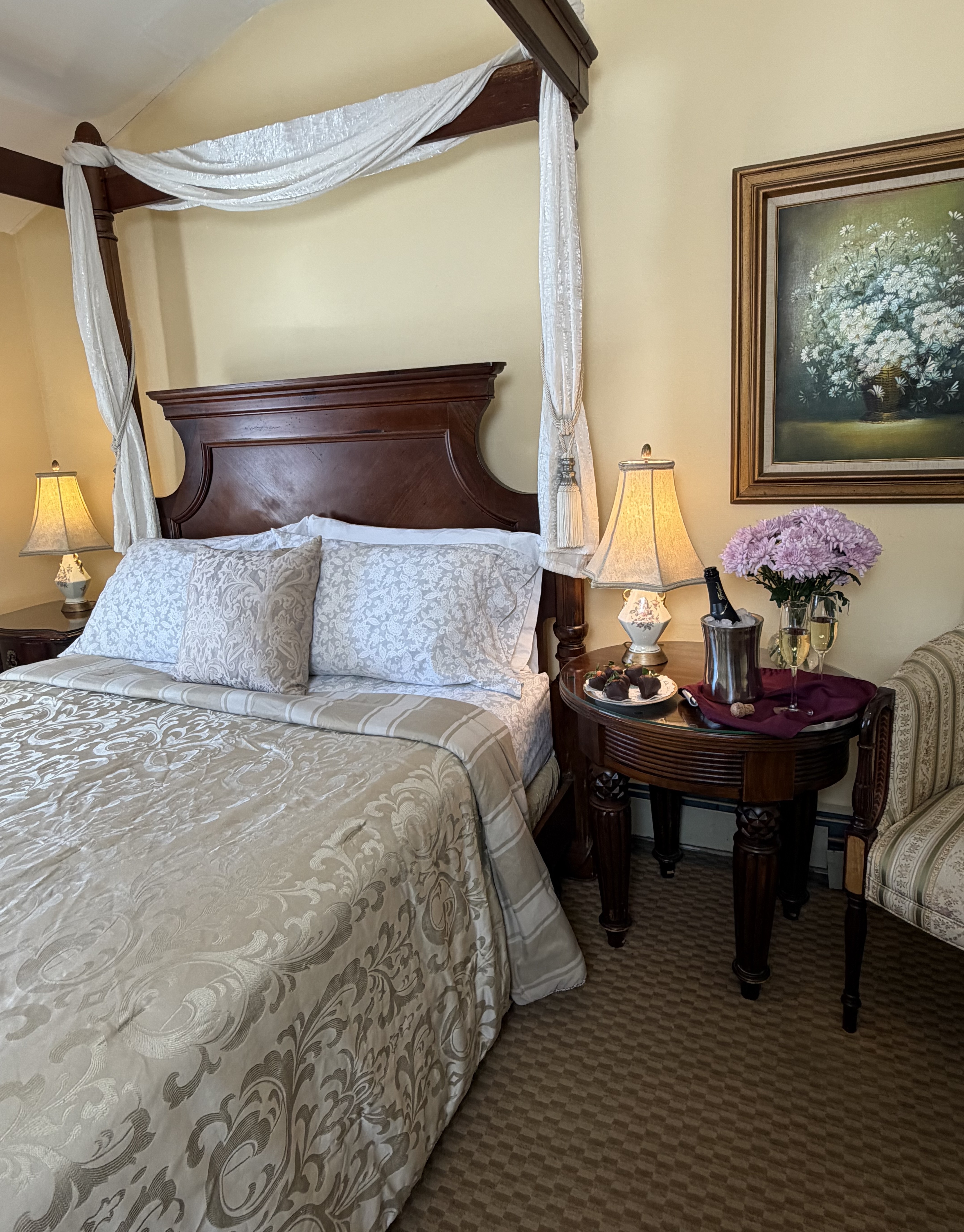 Side view of 4-post walnut queen bed with side table & chair, champagne & flowers