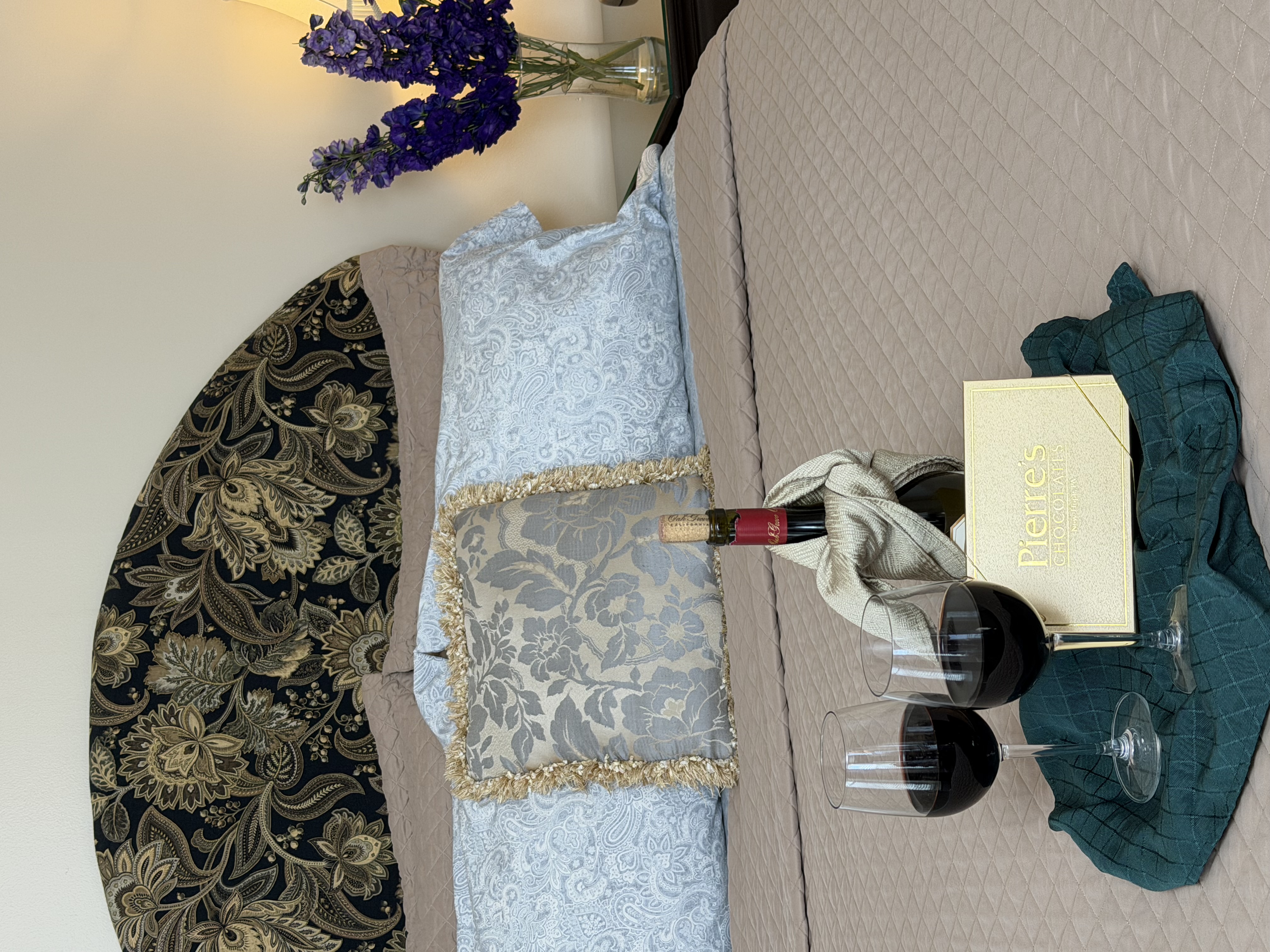 View of queebn bed with flowers on side table, a bottle of read wine with glasses & a box of chocolates