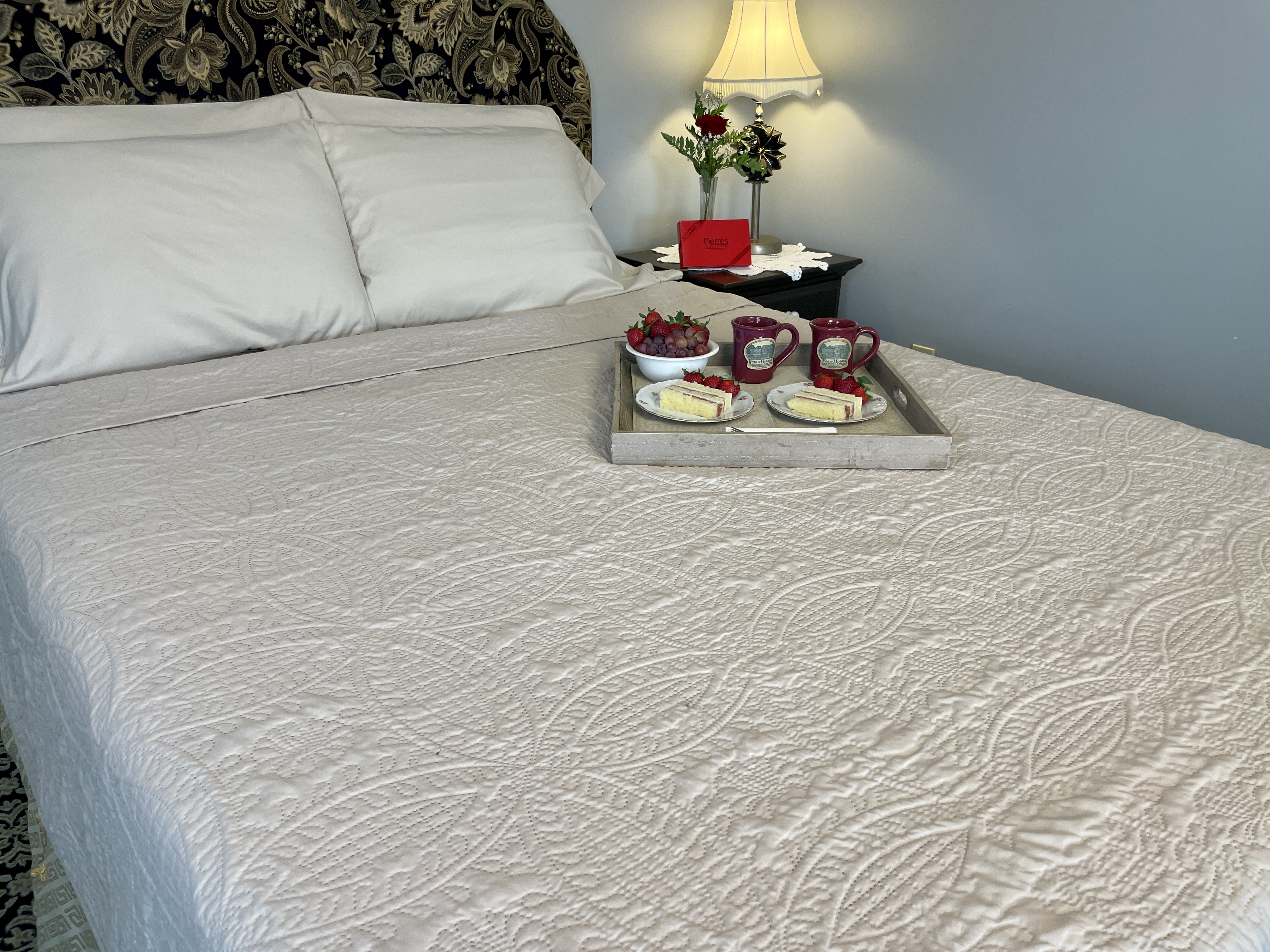 Queen bed with tan coverlet and tray of cakes and coffee on bed with red rose in a vase