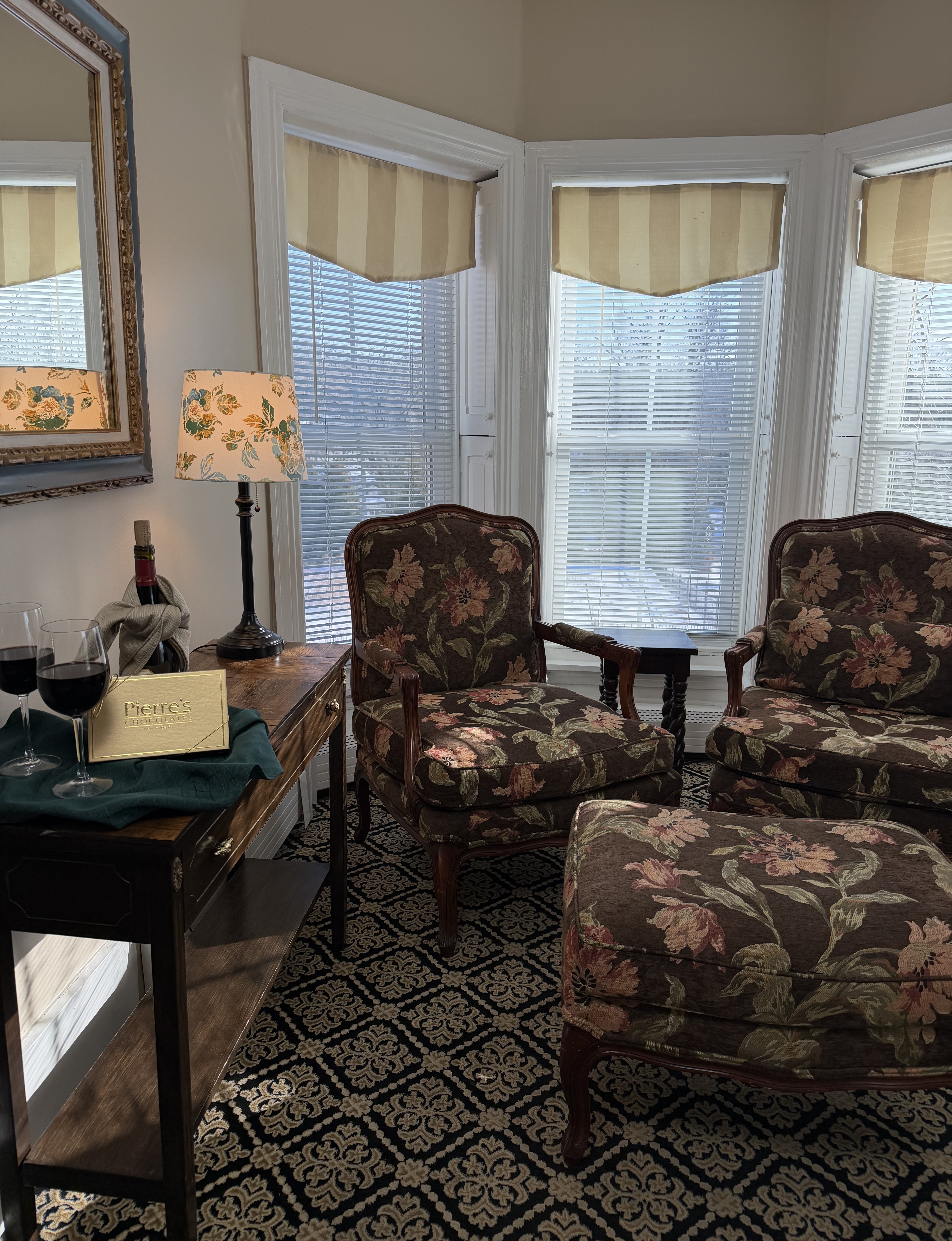 View of bay windows with sitting are, side table, red wine, glasses & a box of chocolates