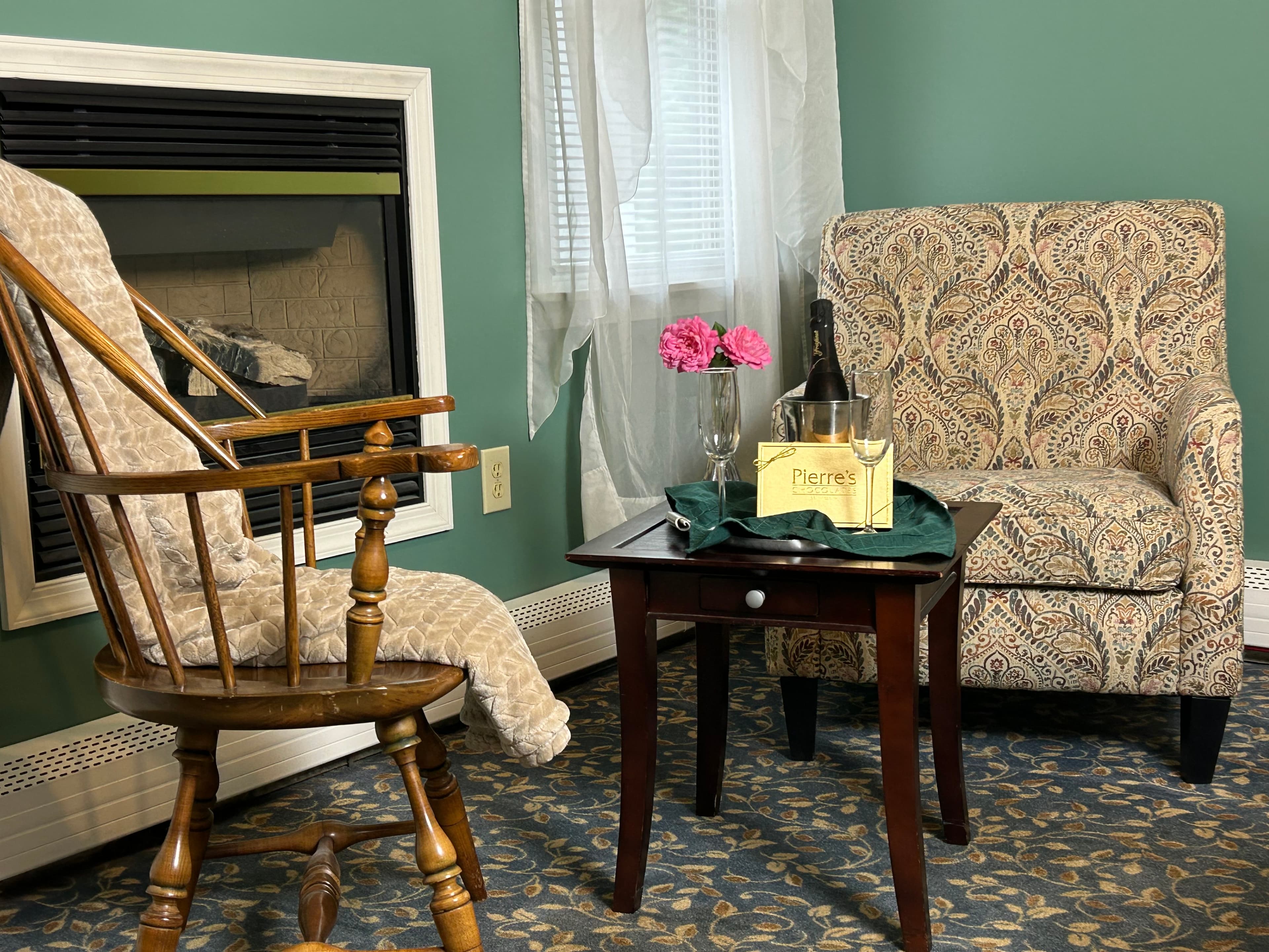View of sitting area and ire place with 2 chairs, table, champagne, 2 glasses and box of chocolates