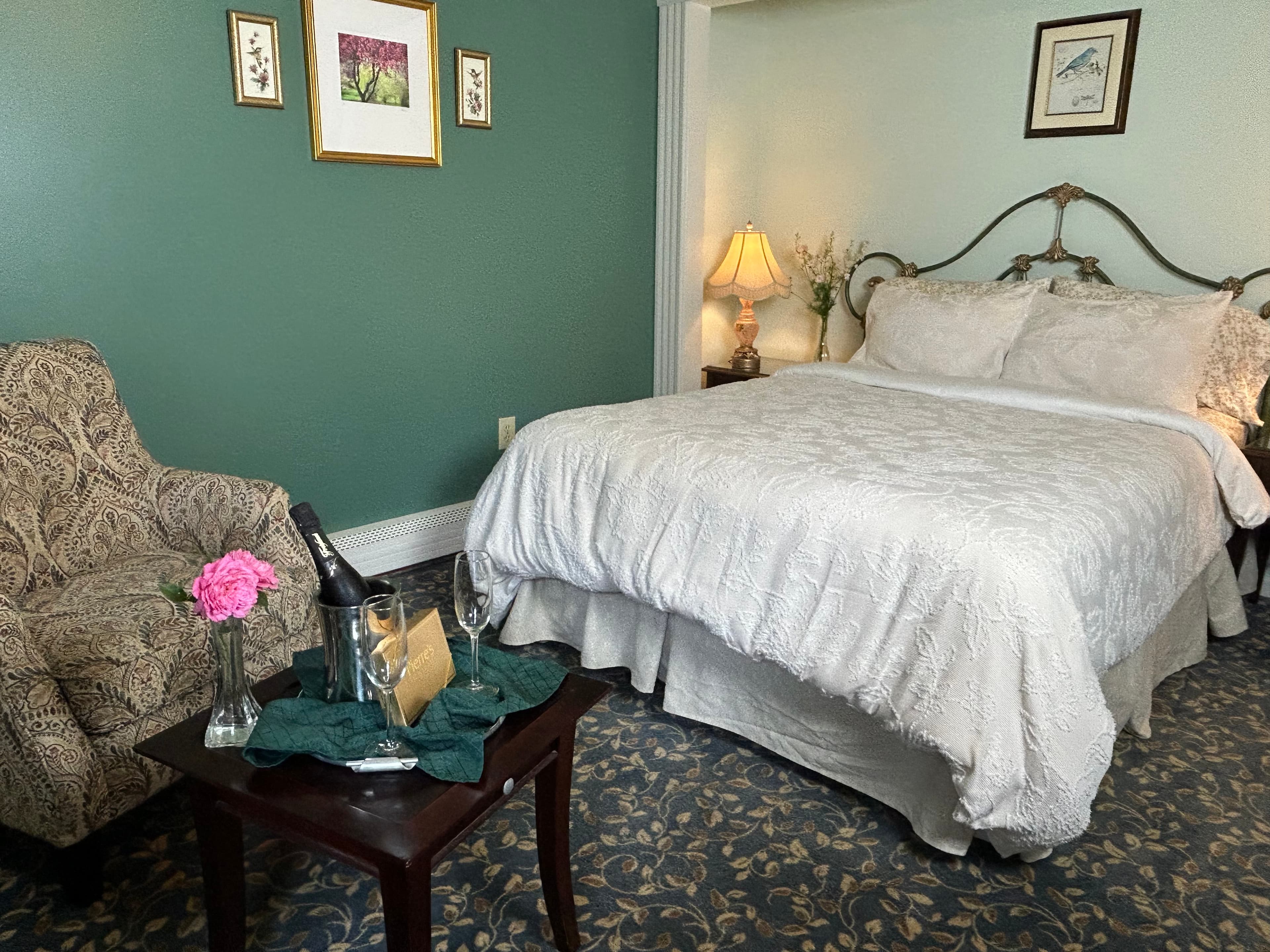 View of queen bed and sitting area with a chair, a table, champagne, 2 glasses and chocolates