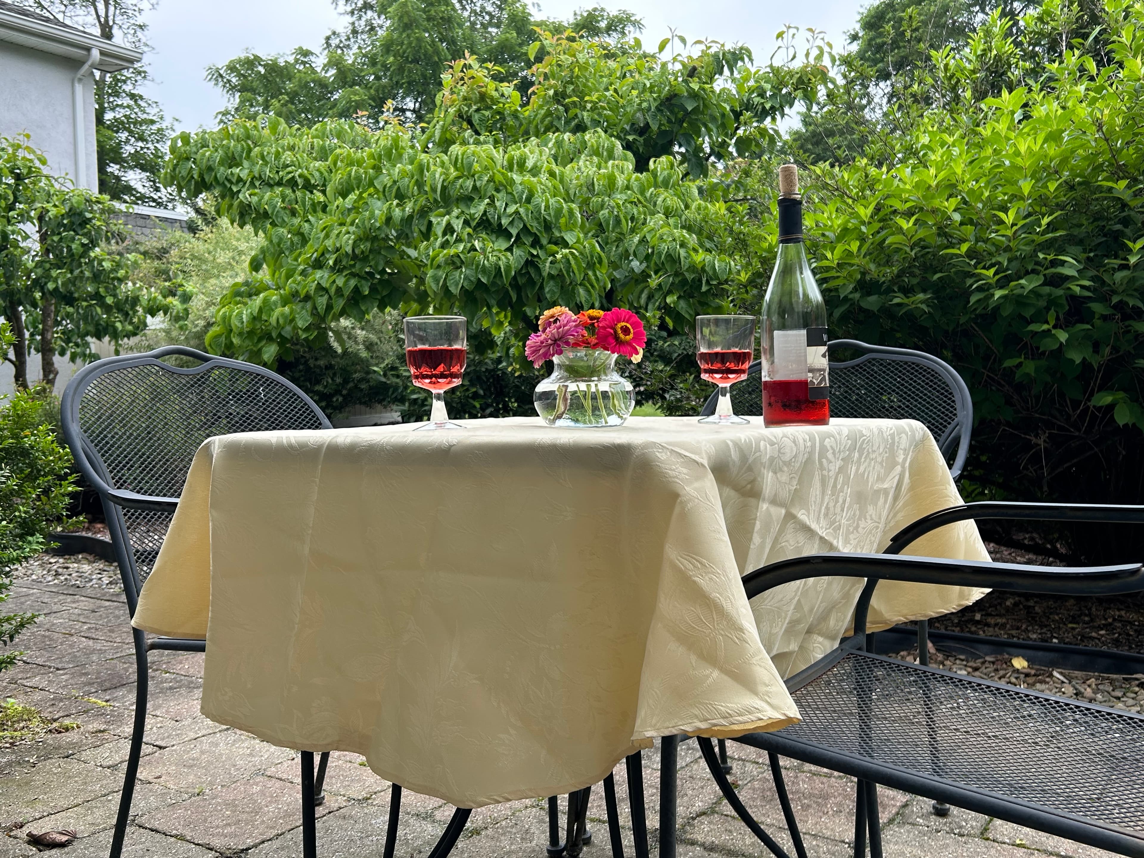 View of back patio with table and chairs, wine, 2 wine glasses and flowers overlooking rear garden