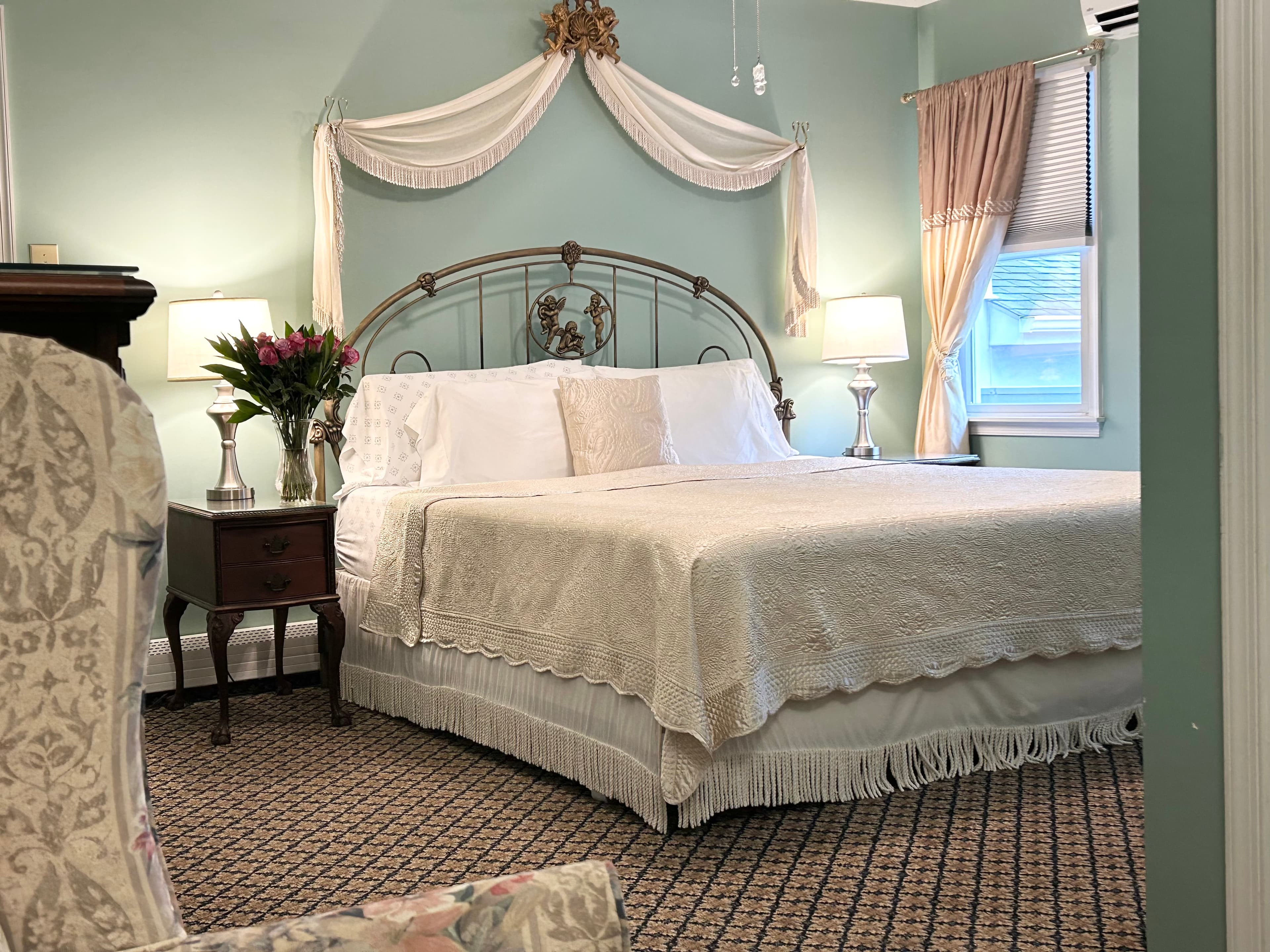 View of king bed with a side table, arm chair, flowers and a window
