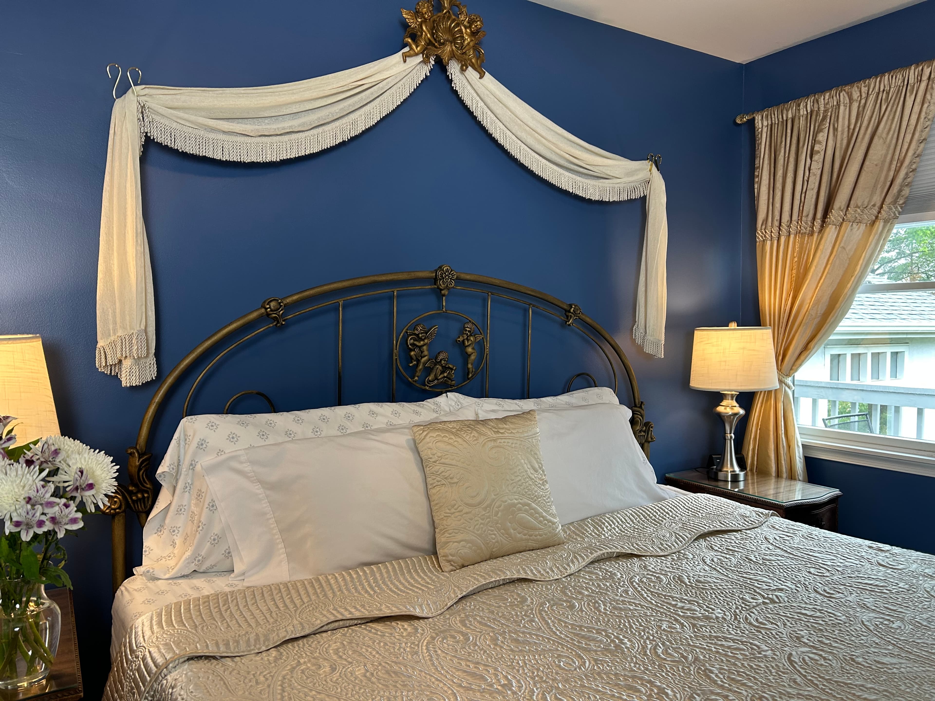 View of king size bed  with swag on wall over the ed, side tables with lamps and a window