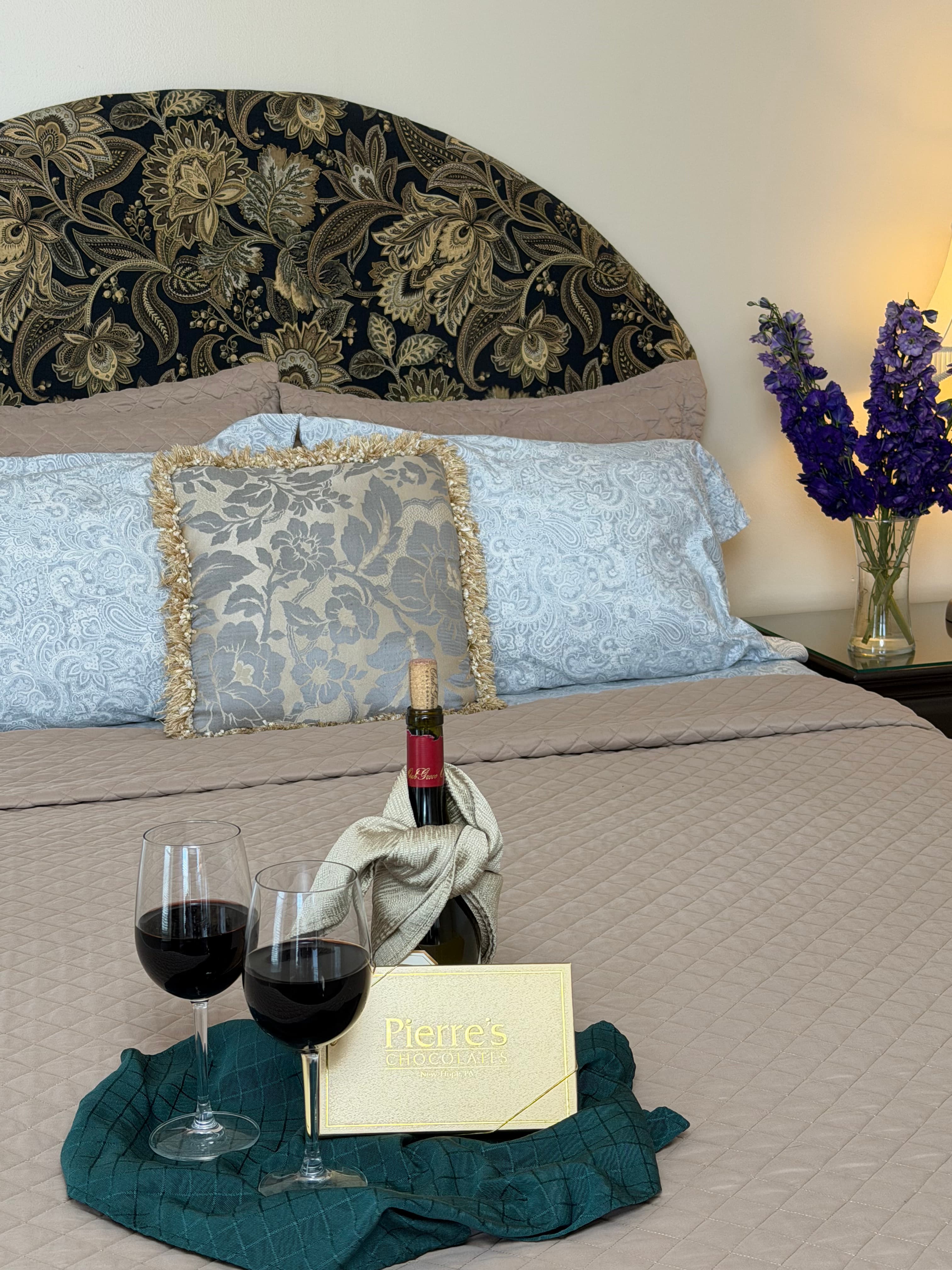 View of queebn bed with flowers on side table, a bottle of read wine with glasses & a box of chocolates