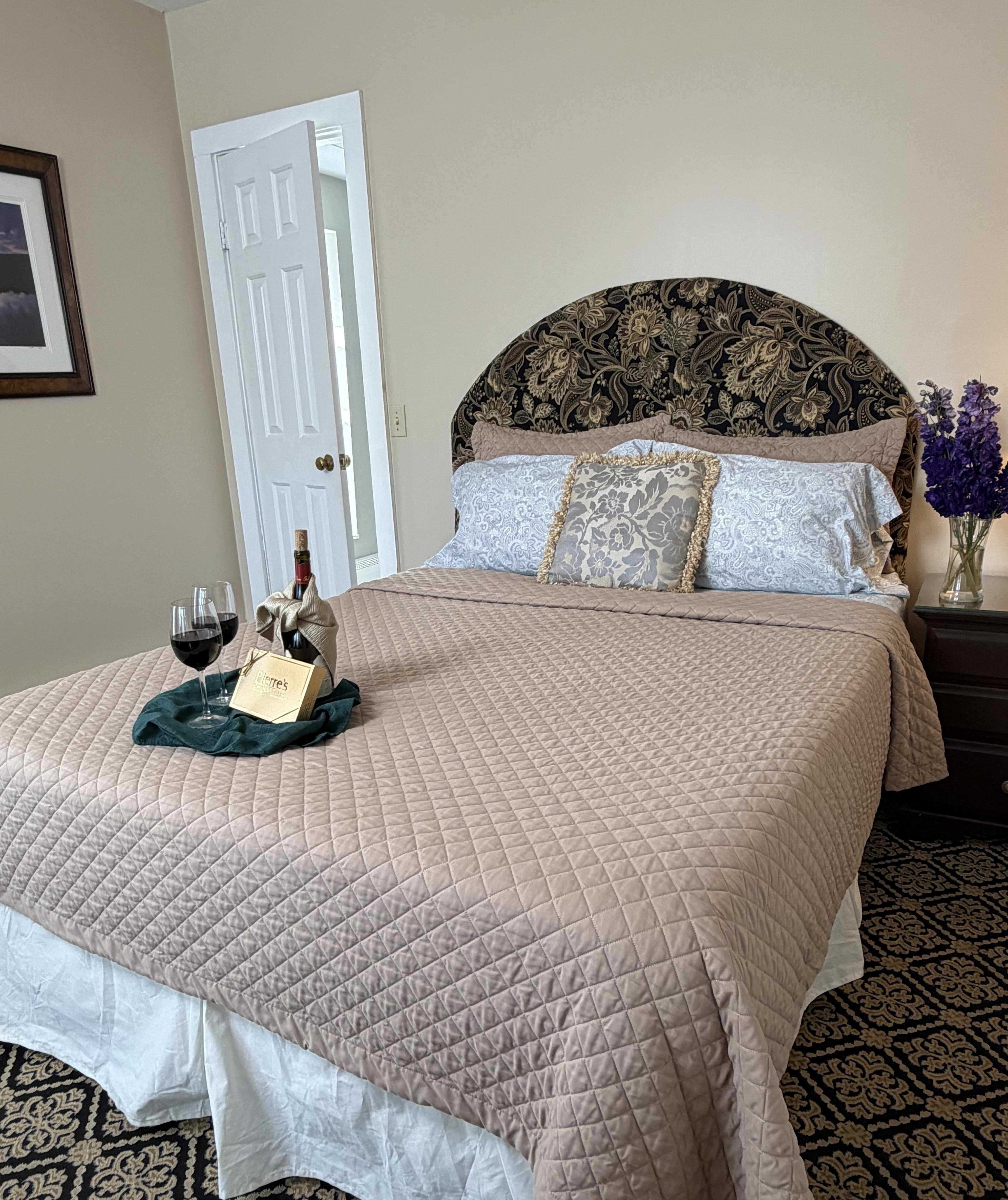 View of queebn bed with flowers on side table, a bottle of read wine with glasses & a box of chocolates, plus door to bathroom