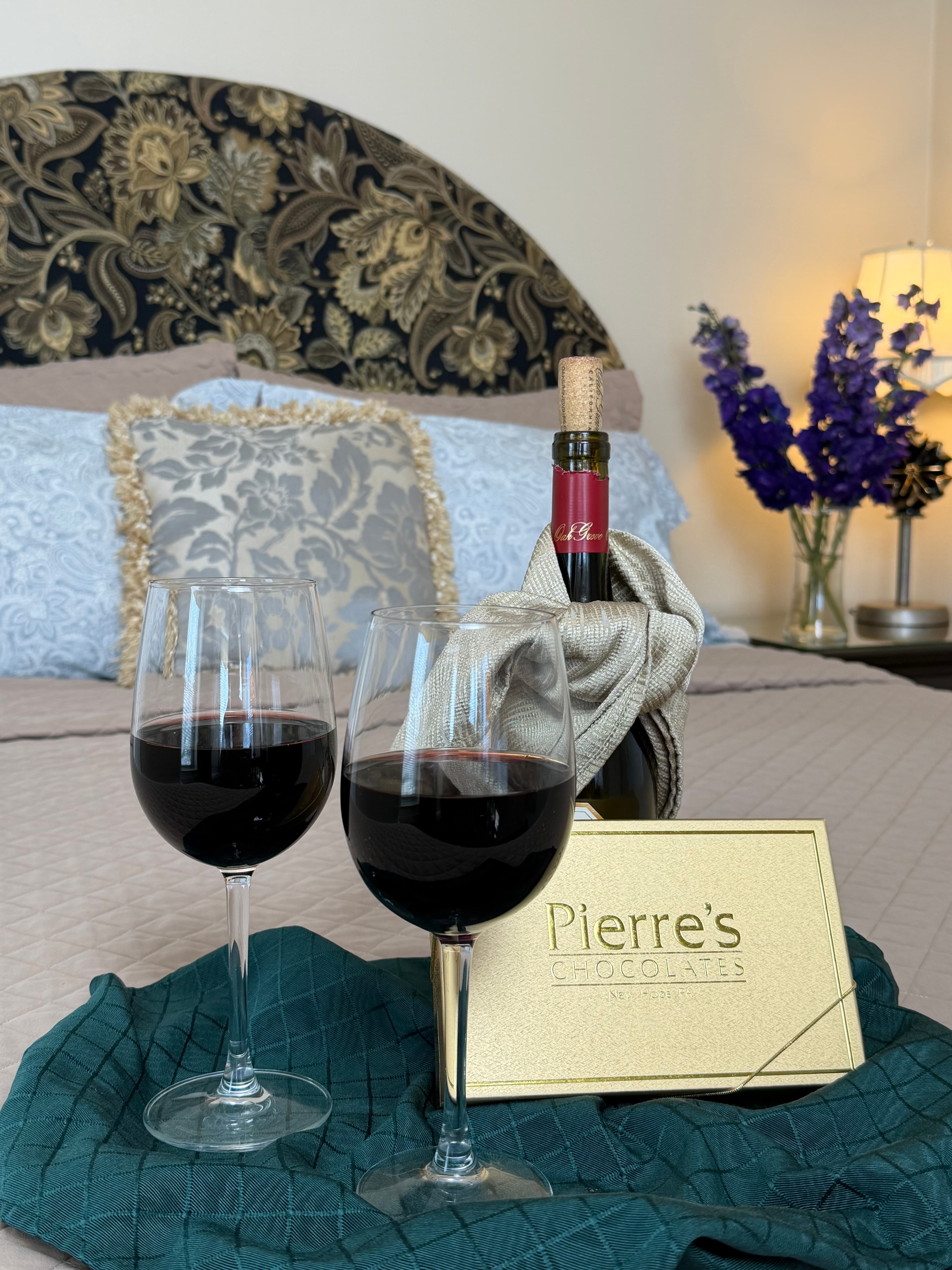 Close up view of a bottle of rd wine, glasses & a box of chocolates on the queen bed