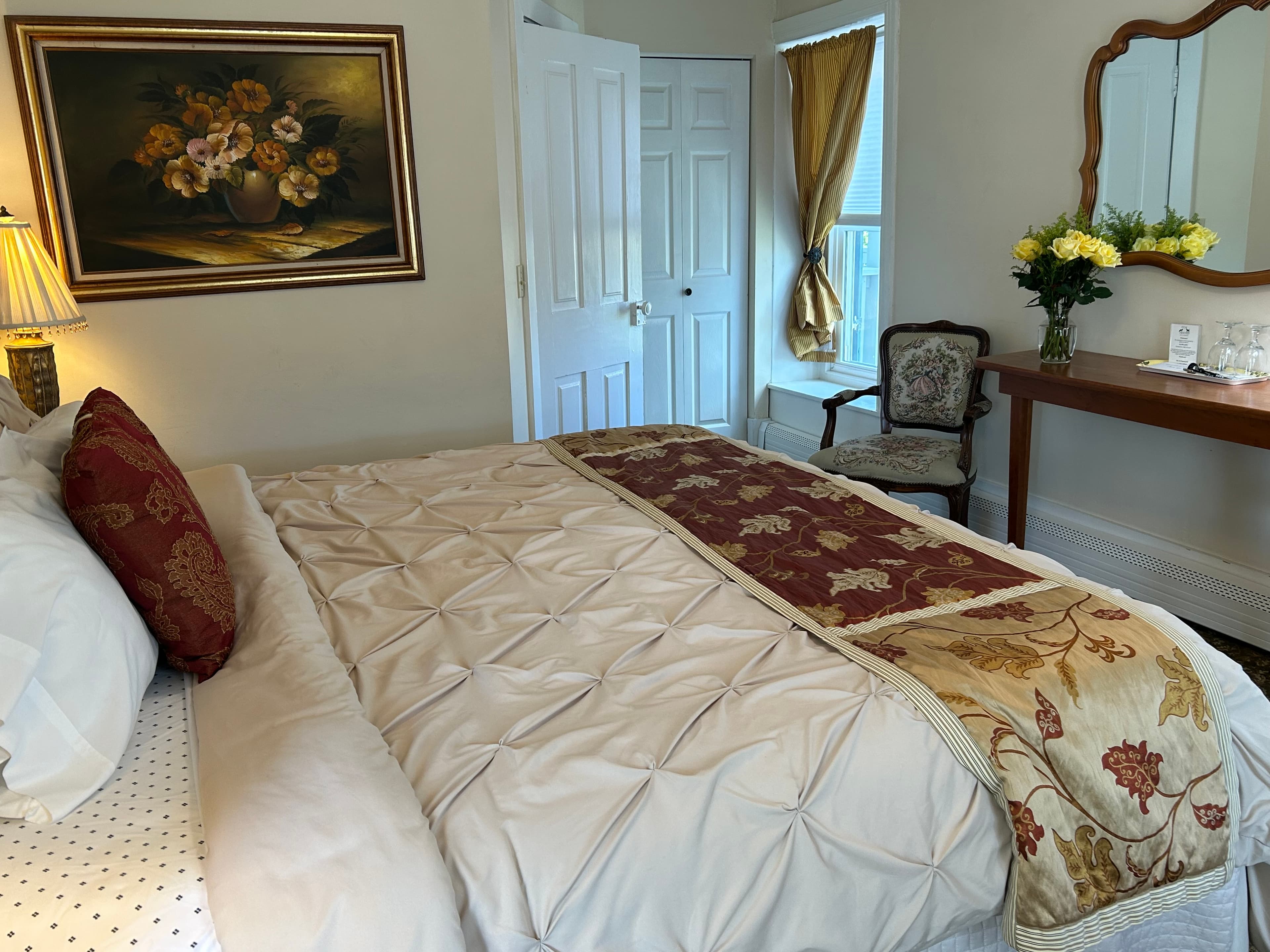 Side view of king bed with painting on the wall, a window, chair, door to bathroom, side table & mirror
