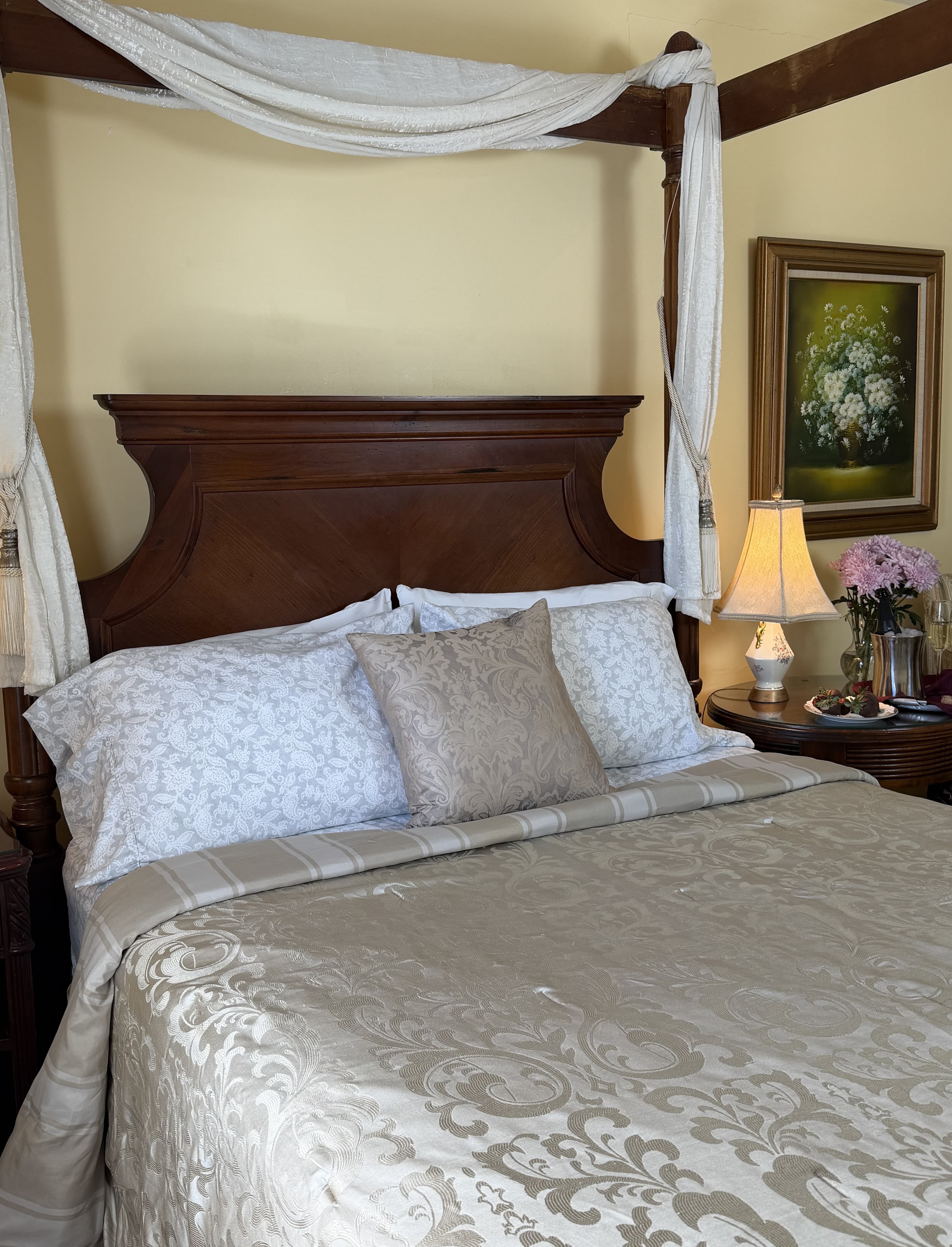 Side view of 4-post walnut queen bed with side table & chair, champagne & flowers