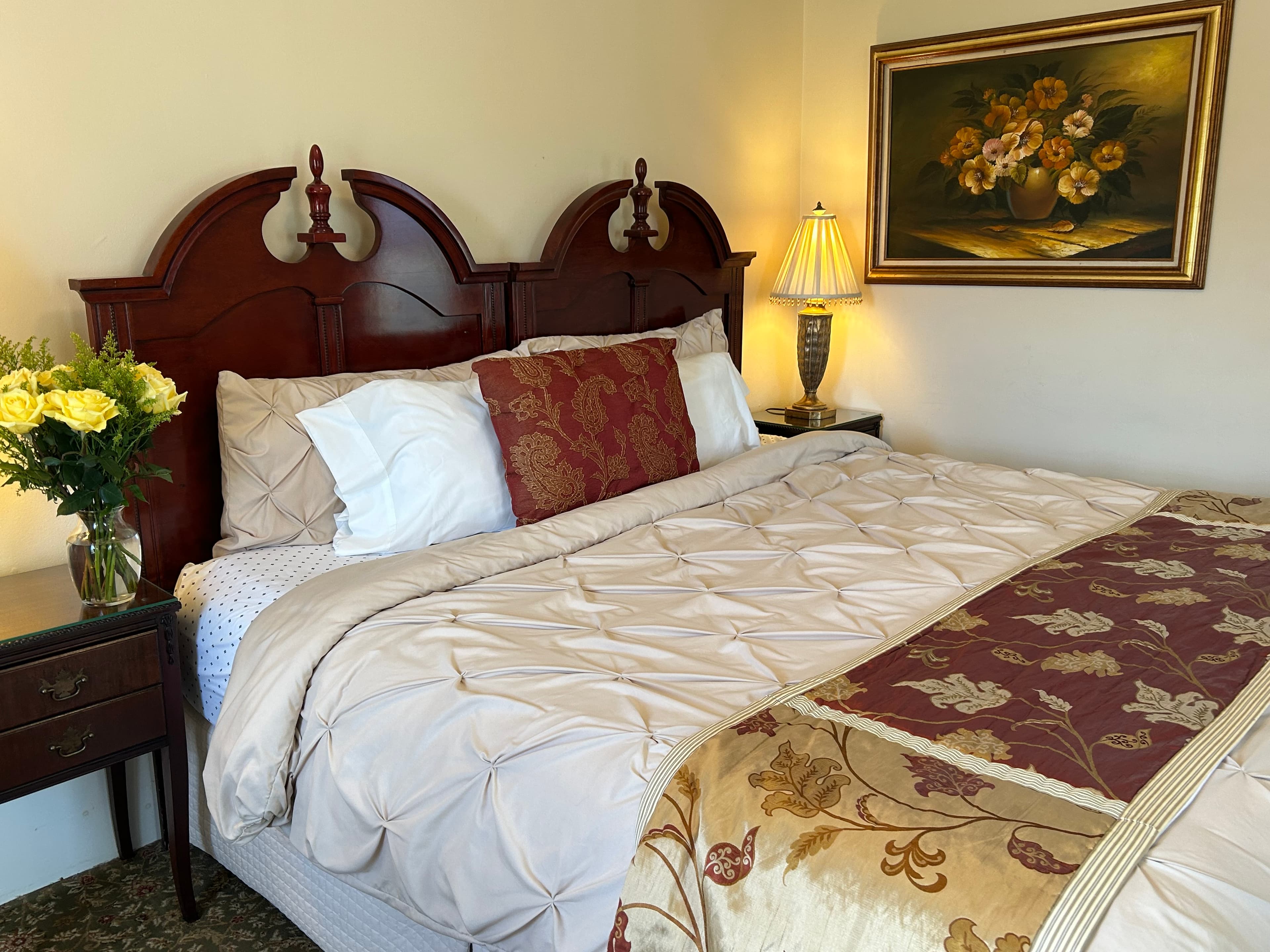 A cozy bedroom featuring a quilted bedspread, decorative pillows, a bedside table with flowers, and a framed painting.