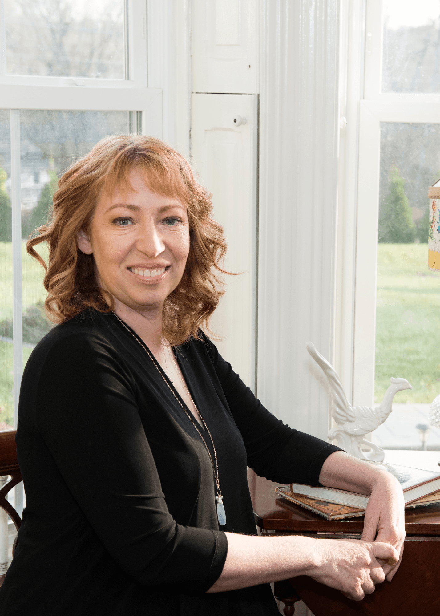 A smiling woman with wavy hair sits by a window, resting her arms on a table with books.