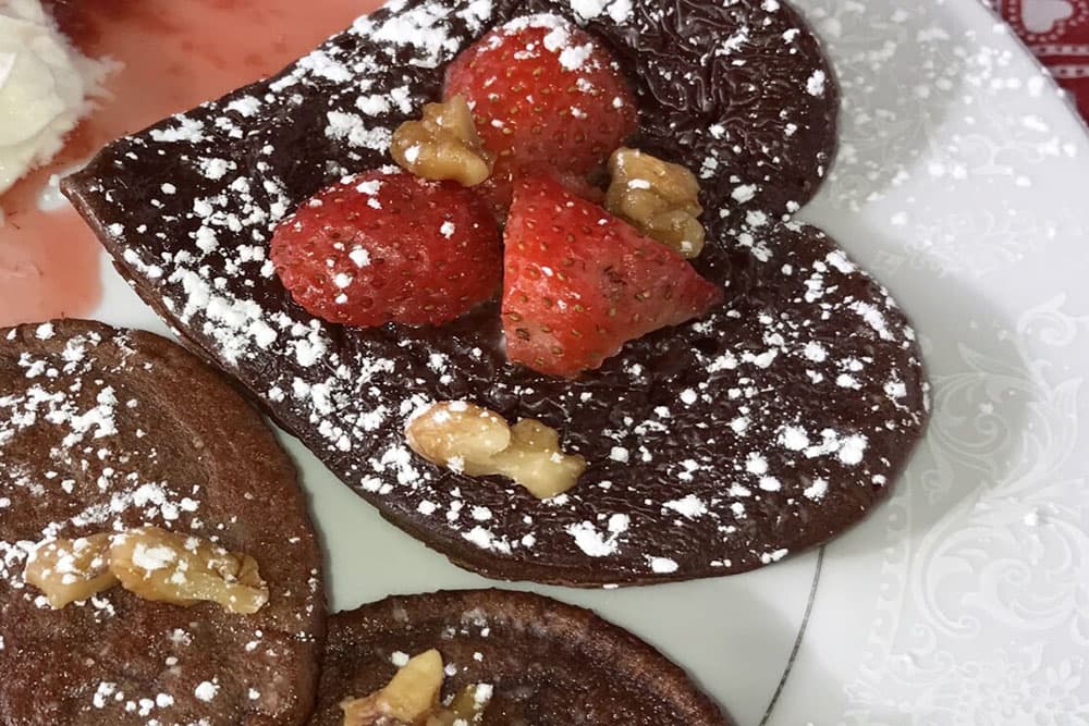 Heart-shaped chocolate pancakes topped with strawberries and walnuts, dusted with powdered sugar.