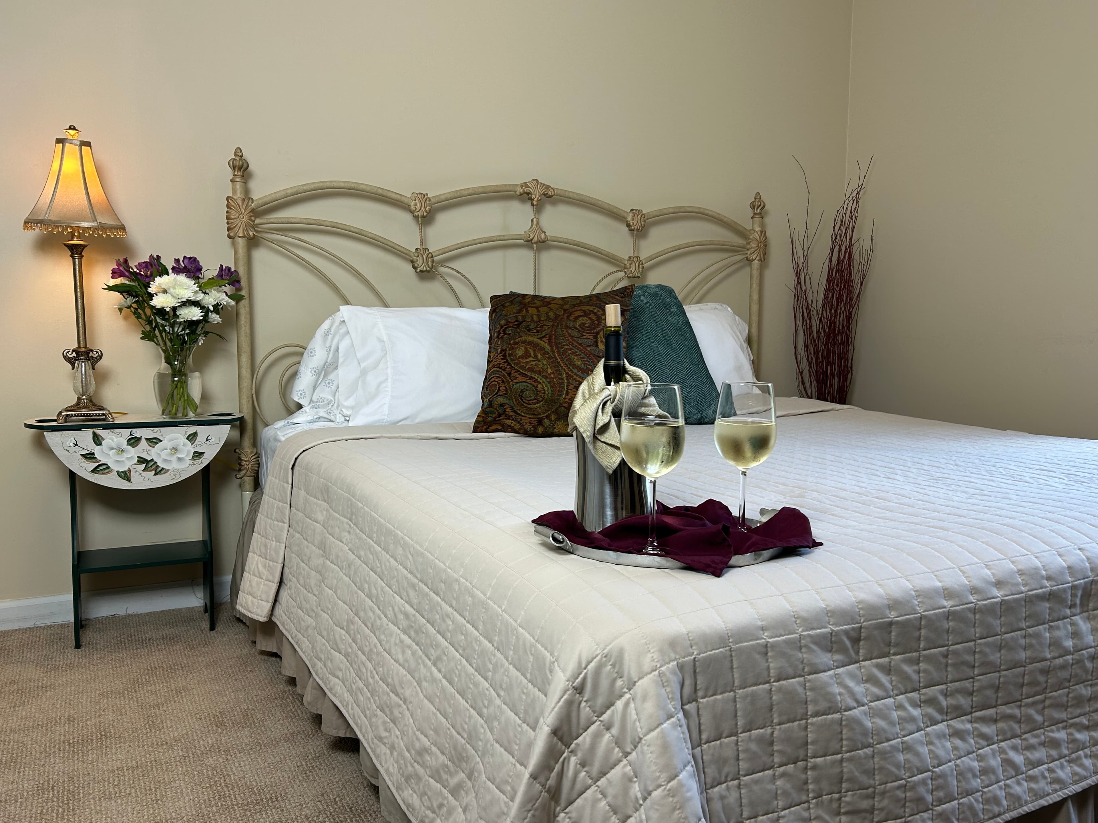 View of king bed, side table & lamp with wine & wine glasses on bed