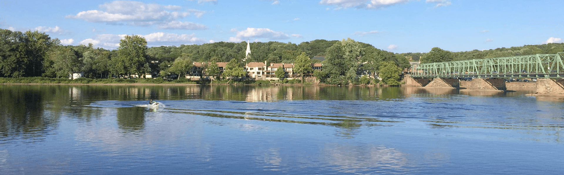 A jet ski creates ripples on a calm river bordered by greenery and buildings under a blue sky.