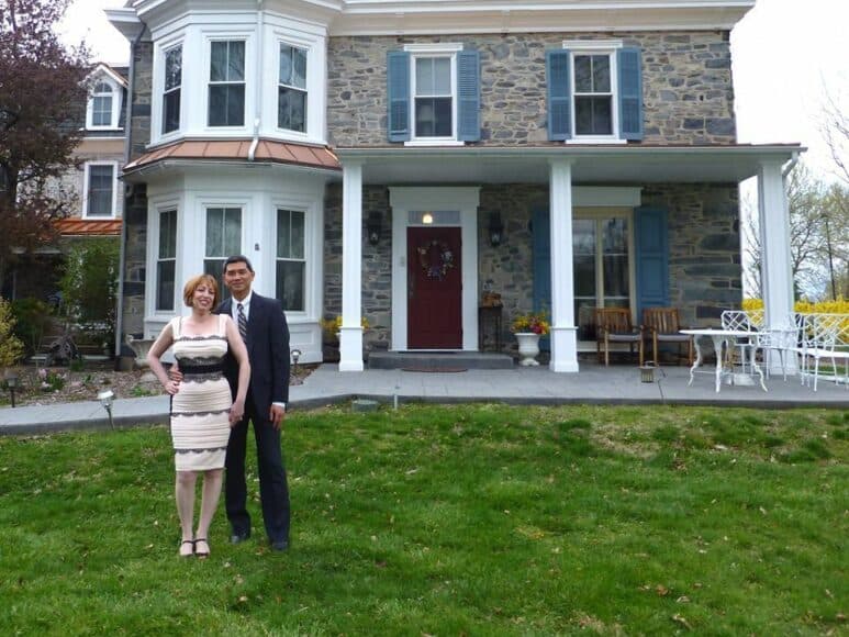 A couple stands together in front of a stone house with blue shutters and a manicured lawn.