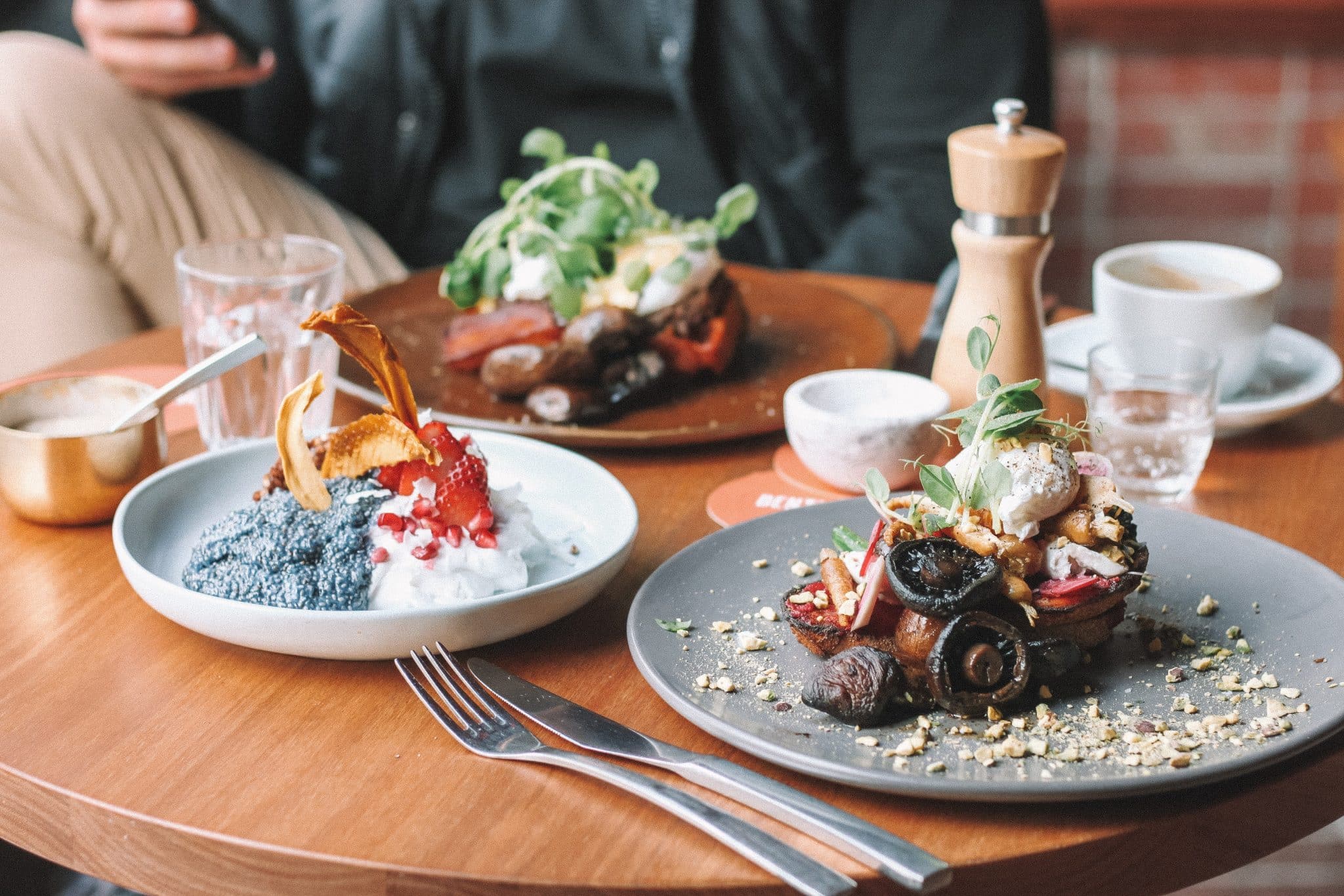 Two beautifully plated dishes on a wooden table, featuring colorful desserts and savory food, accompanied by drinks and utensils.