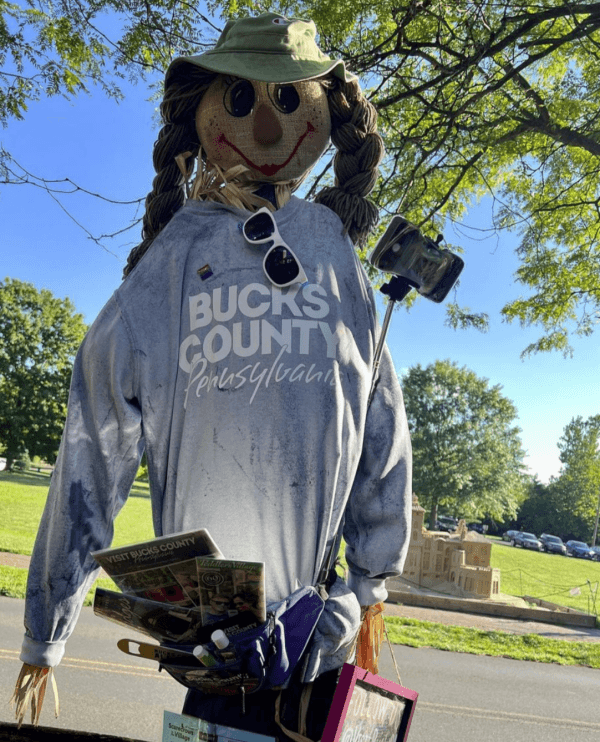 A straw scarecrow wearing a "Bucks County, Pennsylvania" shirt stands outdoors, holding brochures and a camera on a stick.