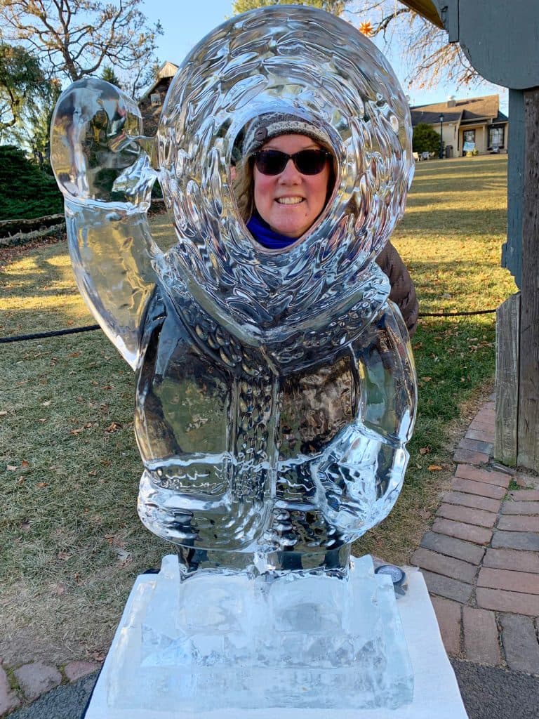 A woman smiles and poses behind a large ice sculpture of a person in a parka waving.