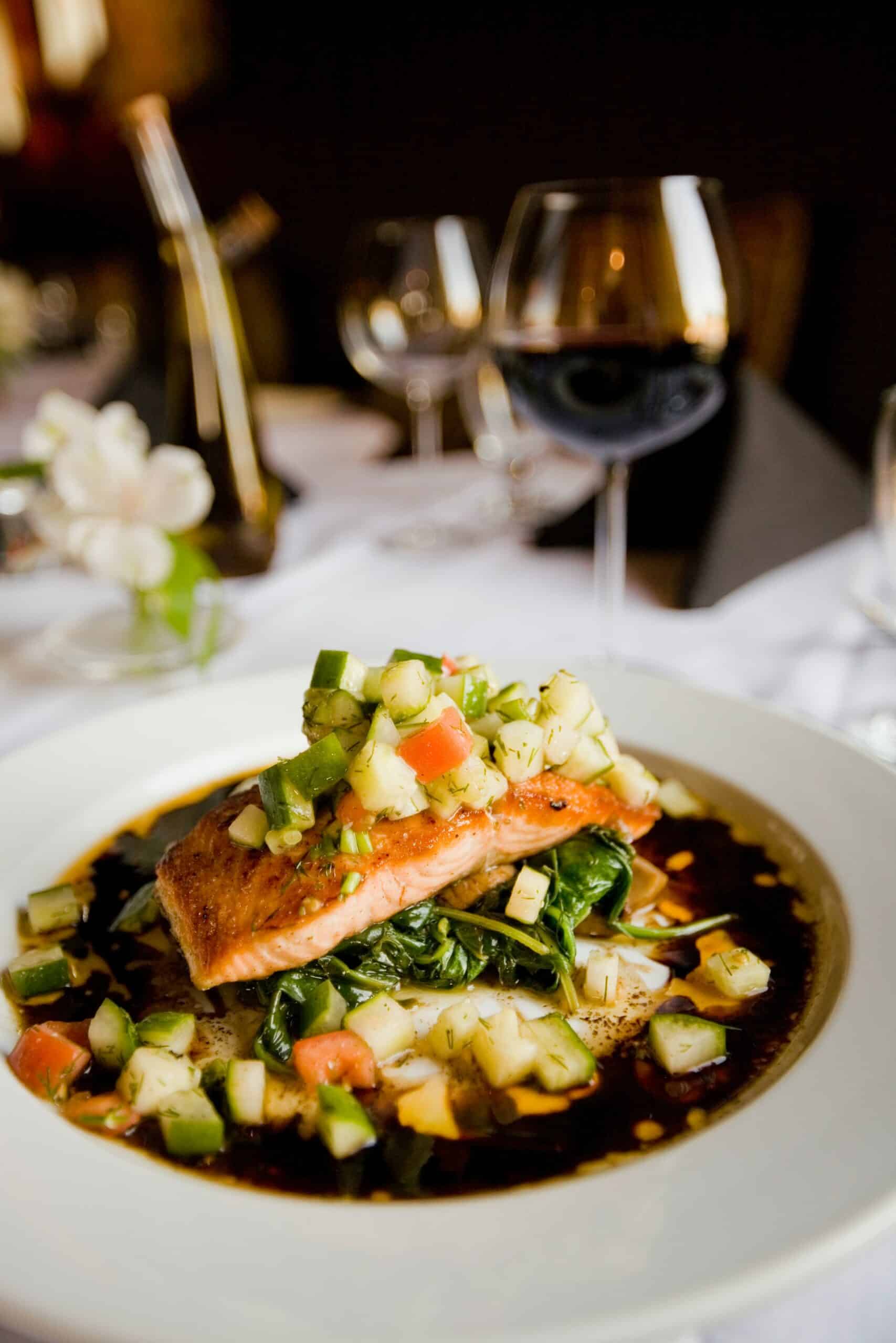 A plated salmon fillet topped with diced vegetables, served on a bed of spinach in a dark sauce.