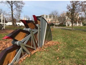 A modern art installation resembling a snow plow rests on a grassy area with a large house in the background.