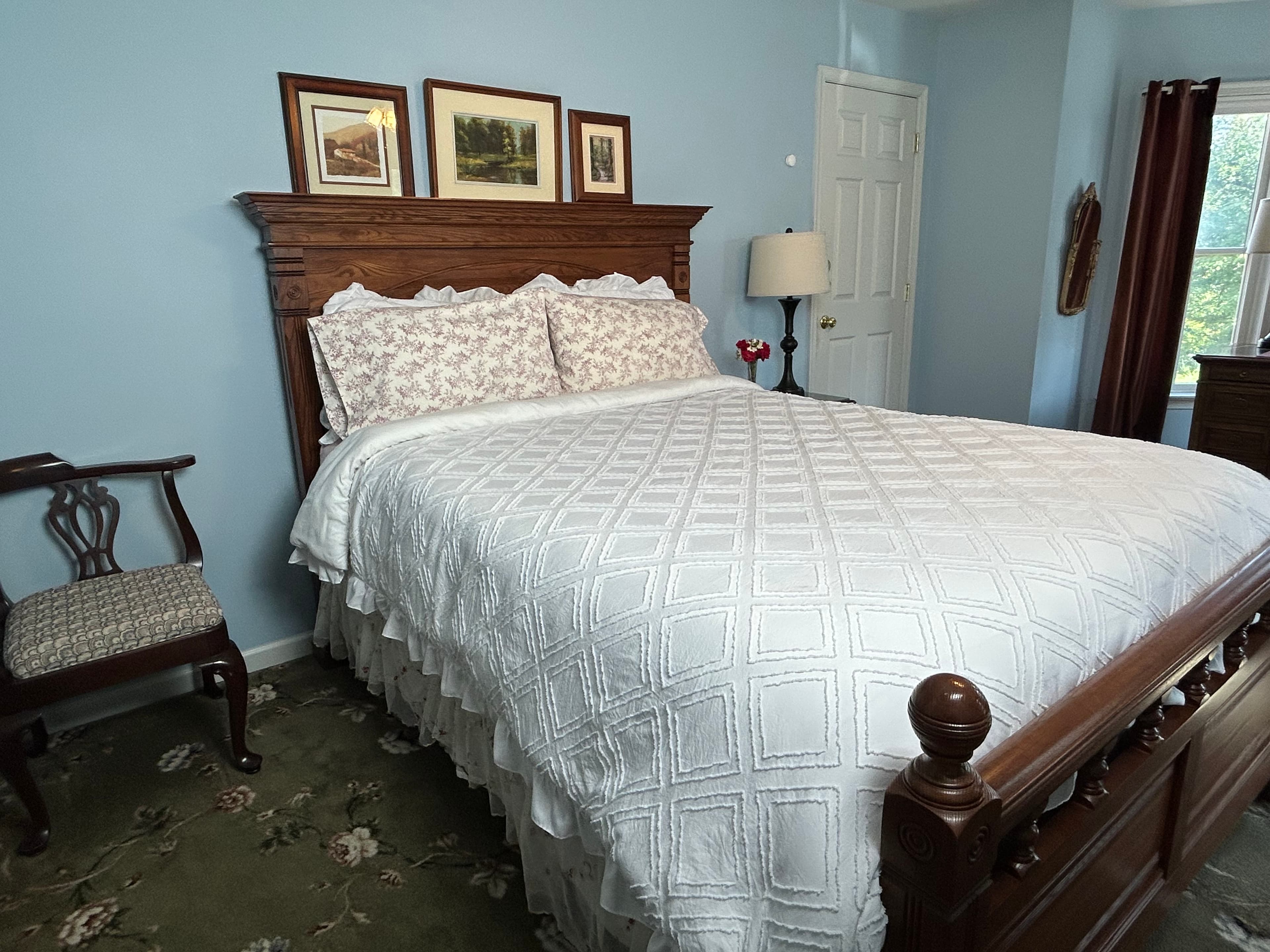View of queen bed with wood headboard, chair, side table with lamp & roses