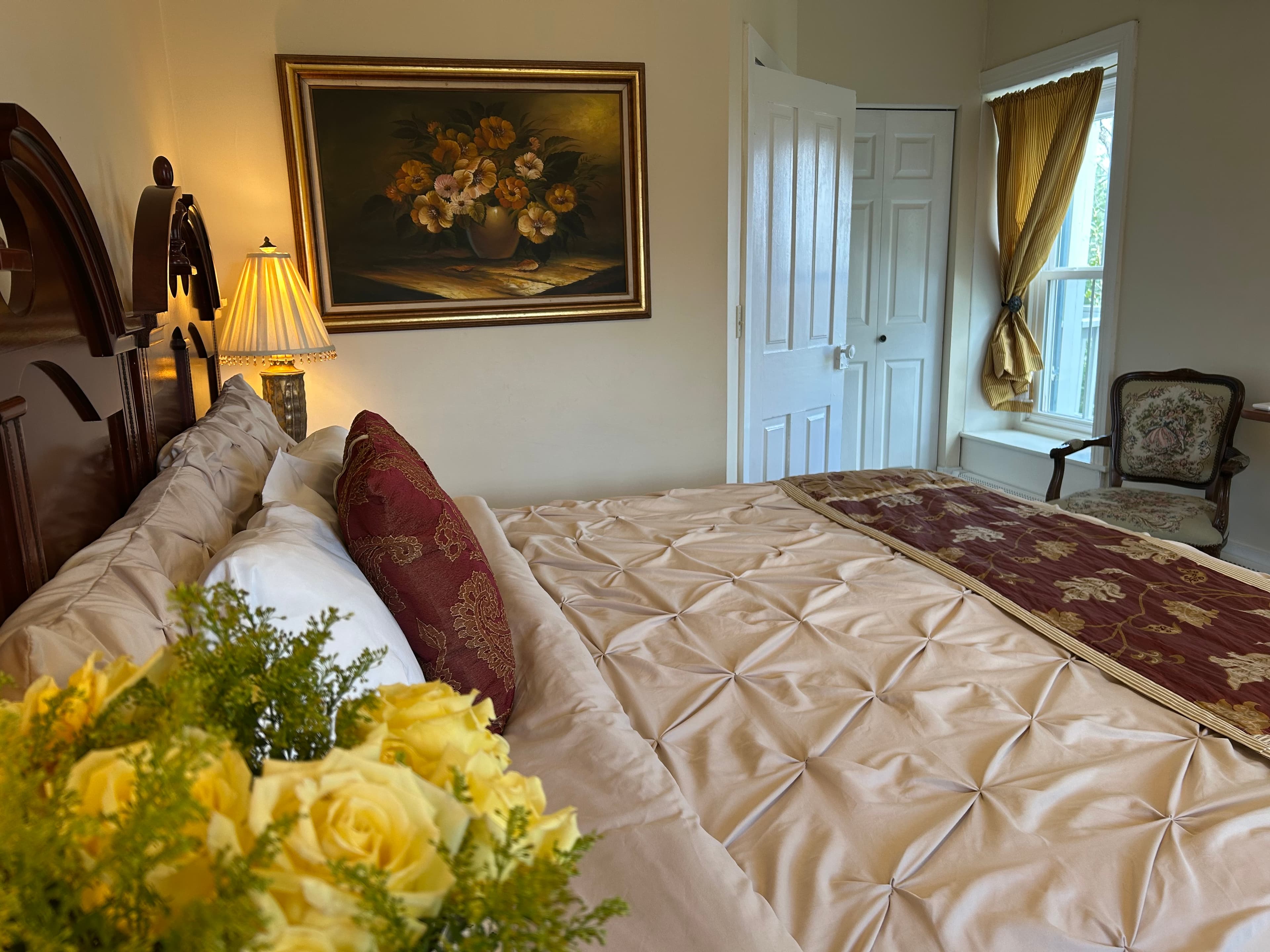 A cozy bedroom featuring a bed with elegant bedding, a bouquet of yellow roses, and a window with drapes.