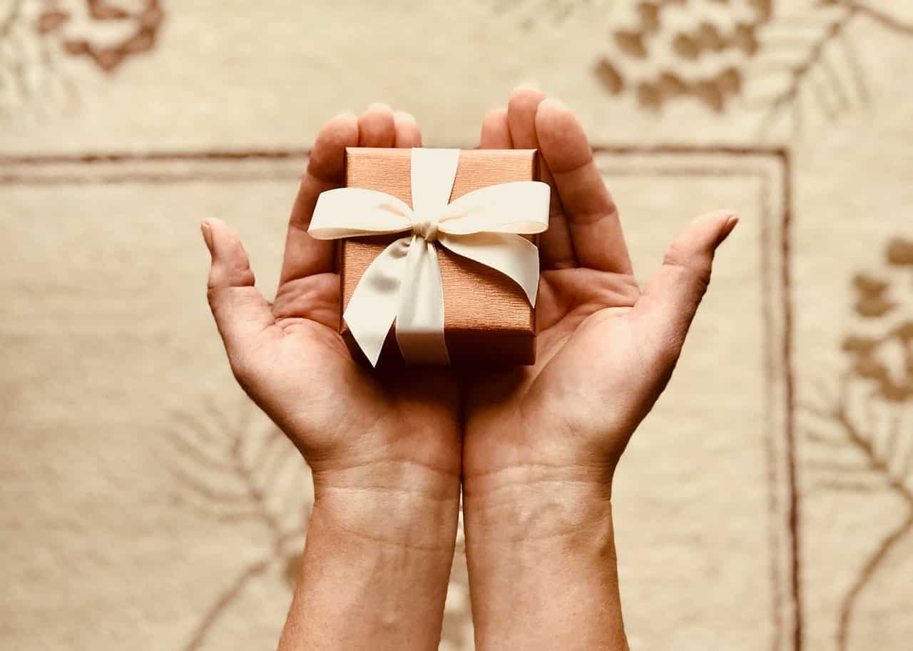 Two hands hold a small, wrapped gift with a ribbon against a patterned background.