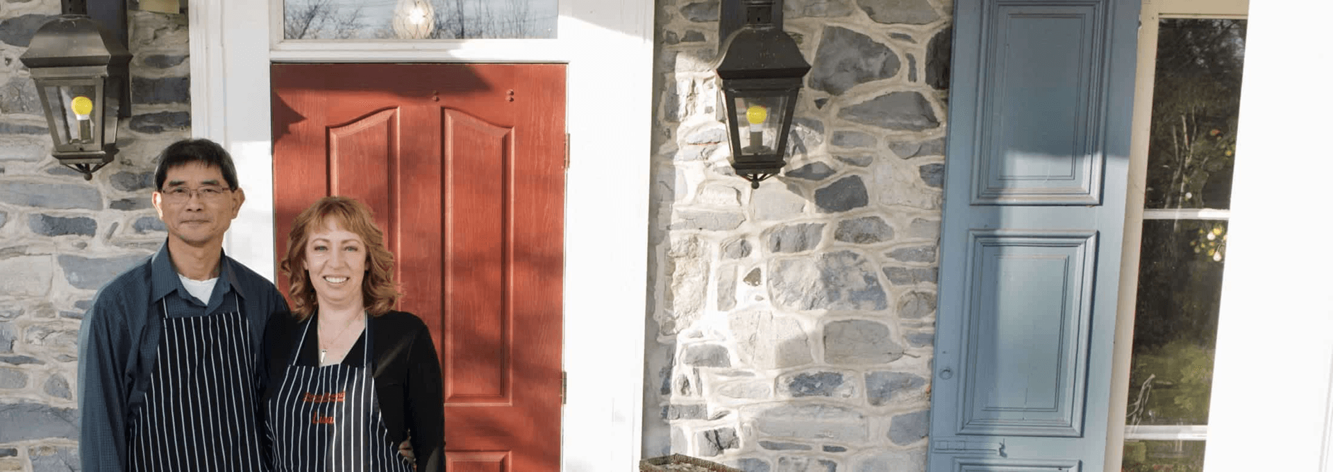 A couple stands together in front of a stone house, both wearing aprons.