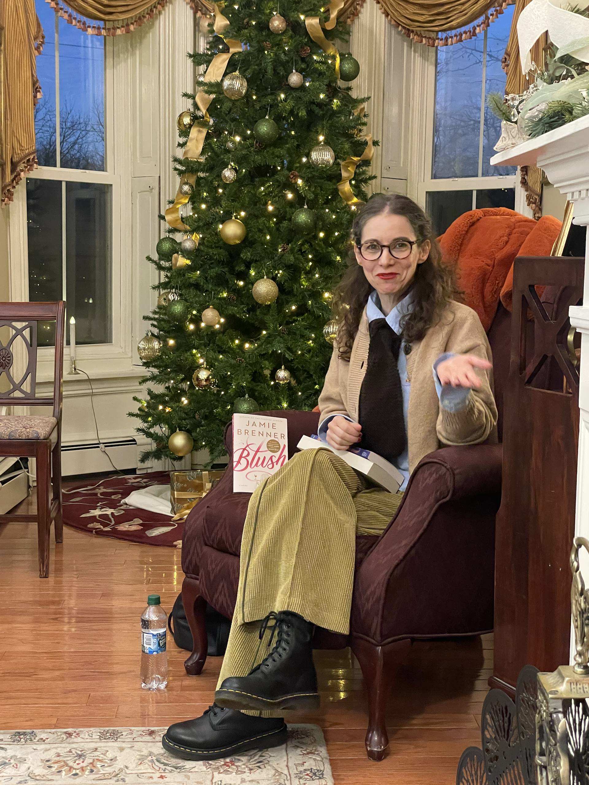 A woman seated in a cozy chair holds a book and gestures, with a decorated Christmas tree in the background.