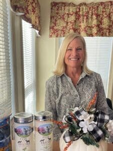 A smiling woman stands beside decorative items, including a pumpkin and two jars, in a sunlit room.