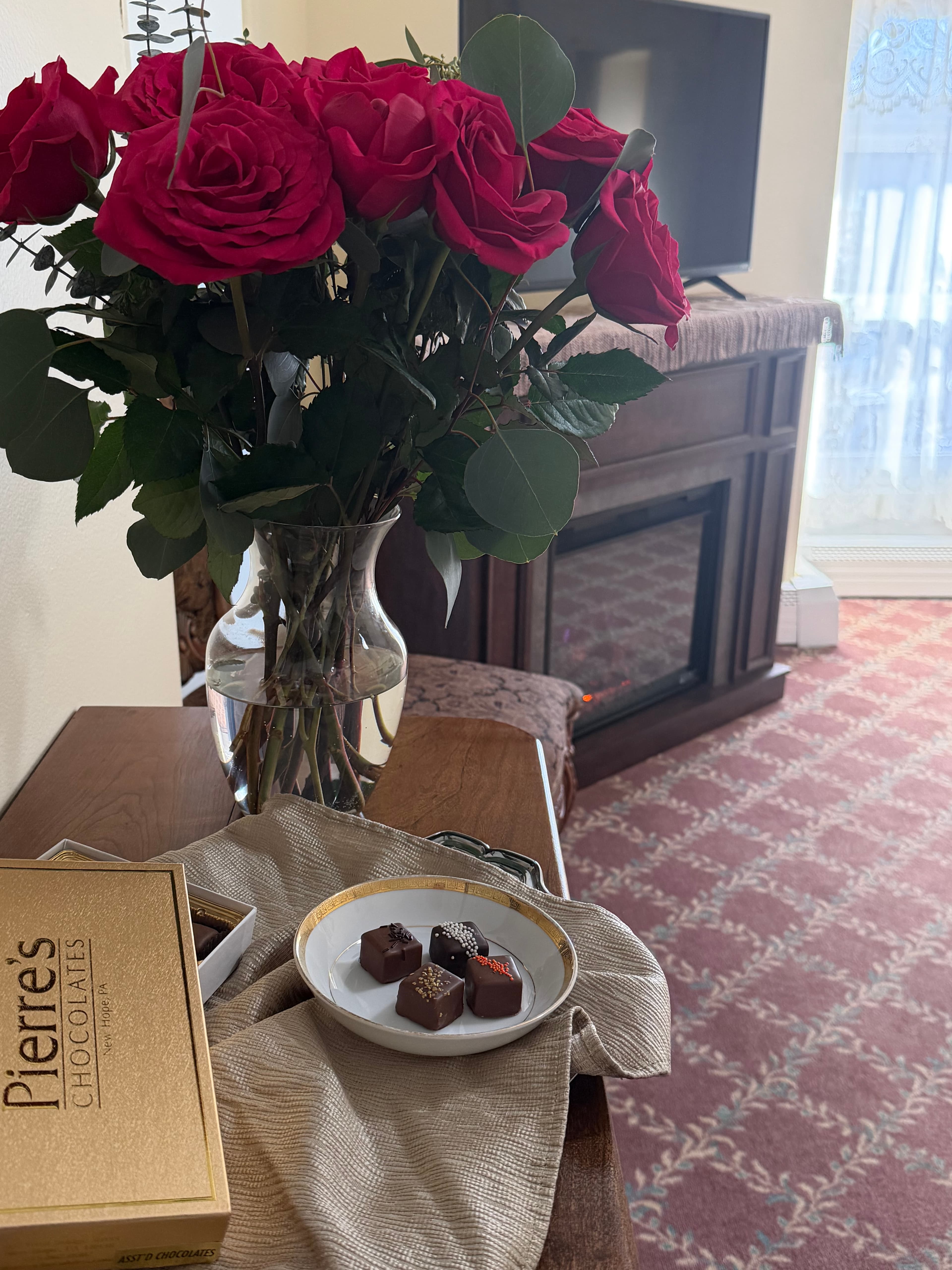 Close up of chocolates & flowers on table by charir, electric fireplace & TV
