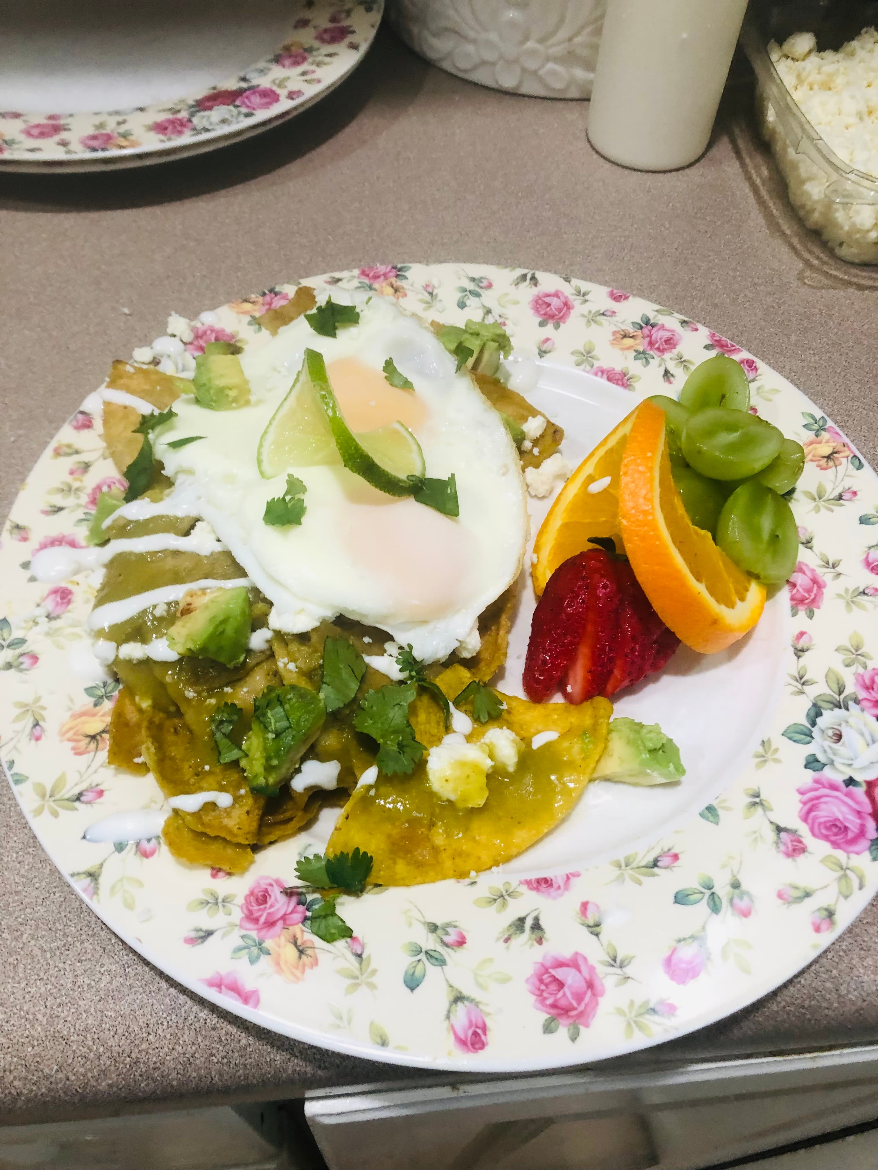 A plate of huevos rancheros topped with a fried egg, avocado, cilantro, and accompanied by assorted fresh fruit.