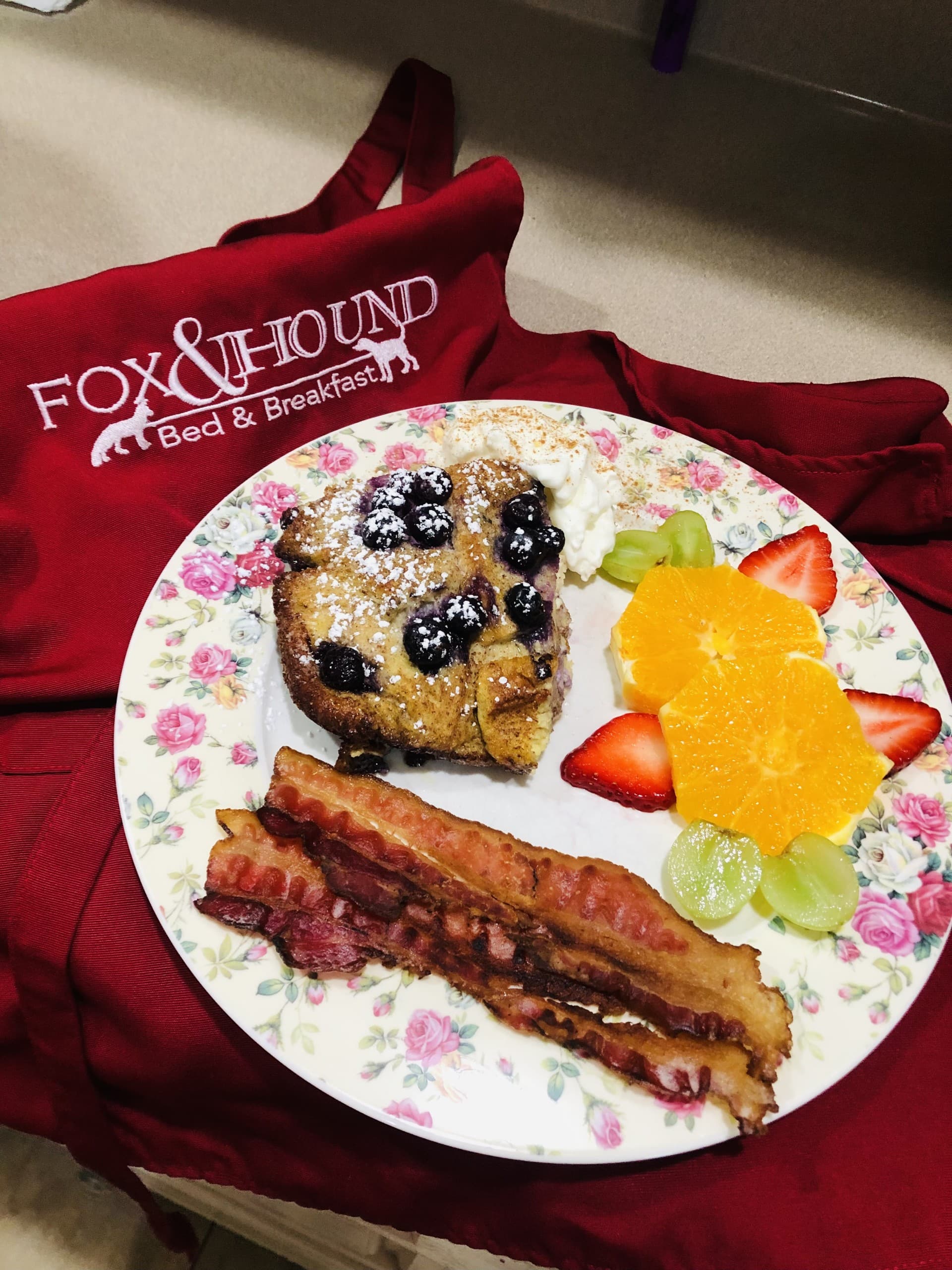 A plate of blueberry French toast, fresh fruit, and crispy bacon on a floral dish, with a red apron in the background.