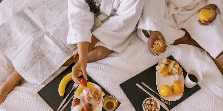 Two people in bathrobes enjoying a breakfast spread on a bed, including fruits and pastries.