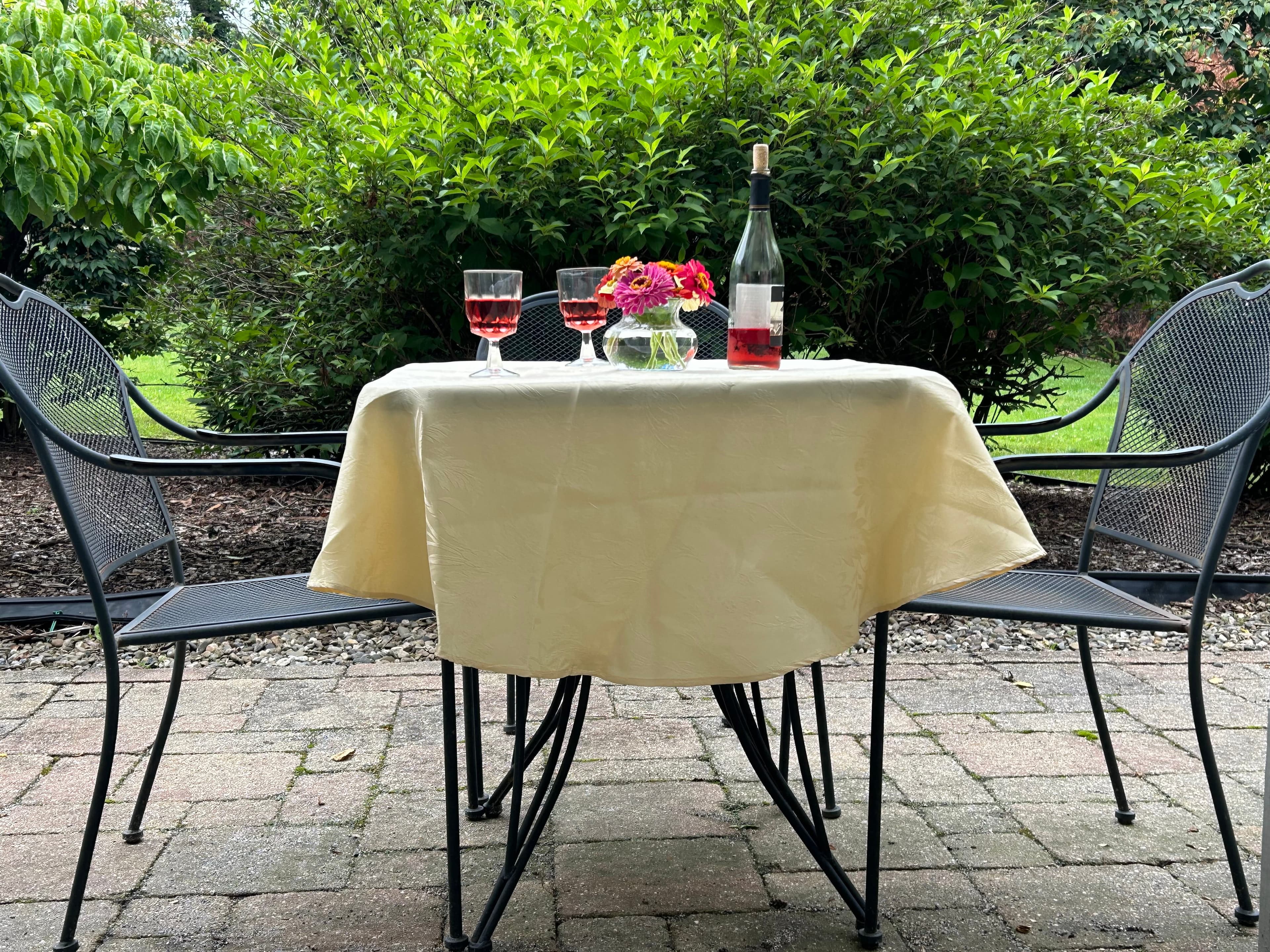 A small outdoor table set with two glasses of pink wine, a flower vase, and a wine bottle, surrounded by chairs and greenery.