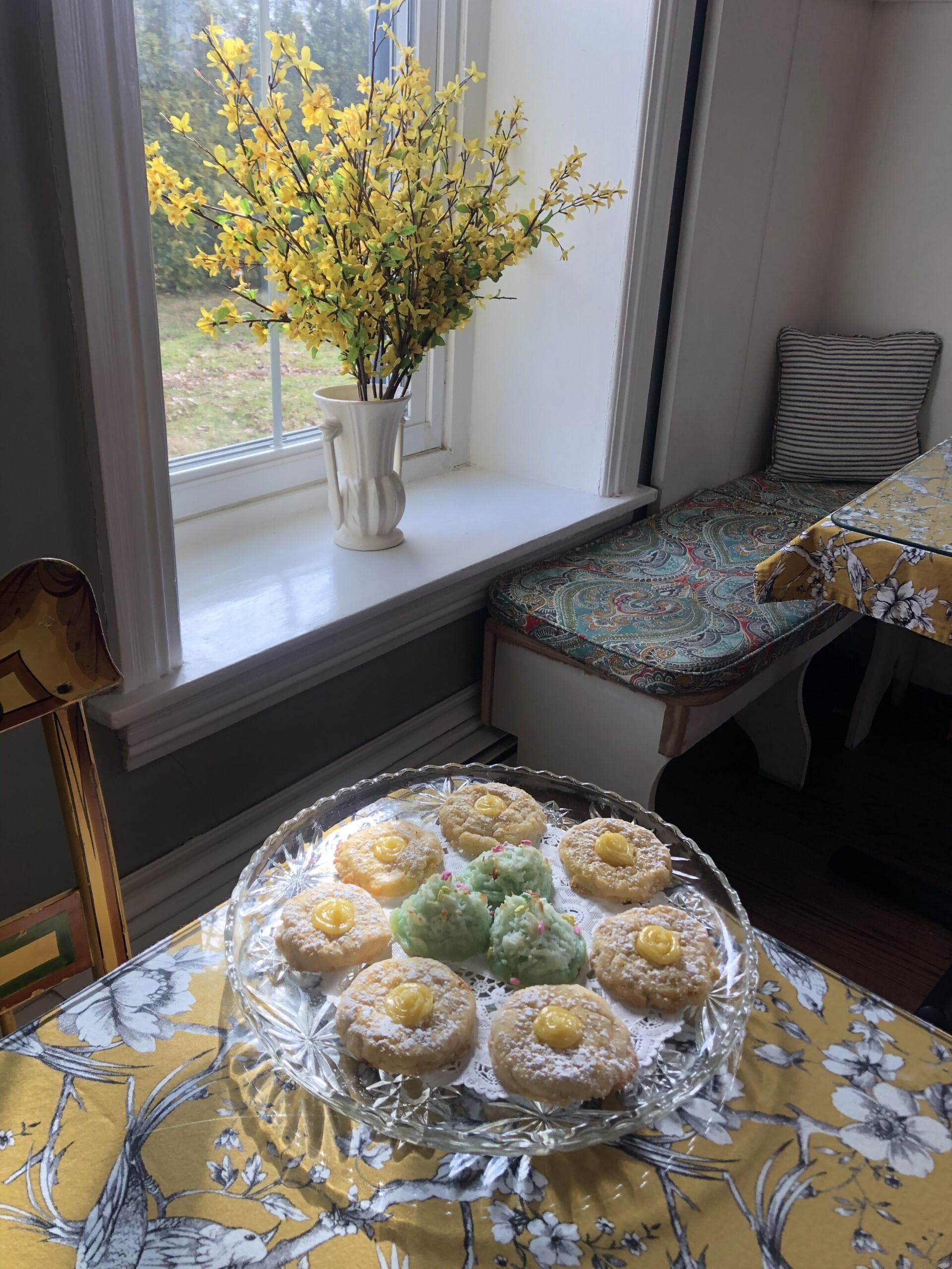 A glass plate of decorated cookies sits on a floral tablecloth by a sunny window with a vase of yellow flowers.