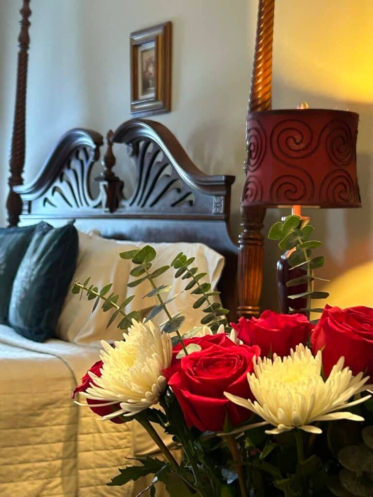 A cozy bedroom featuring a wooden bed frame, decorative lamp, and a bouquet of red roses and white flowers in the foreground.