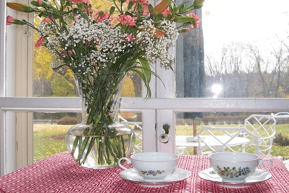 A vase of flowers sits on a table with two delicate tea cups, viewed through a window with an outdoor garden scene.