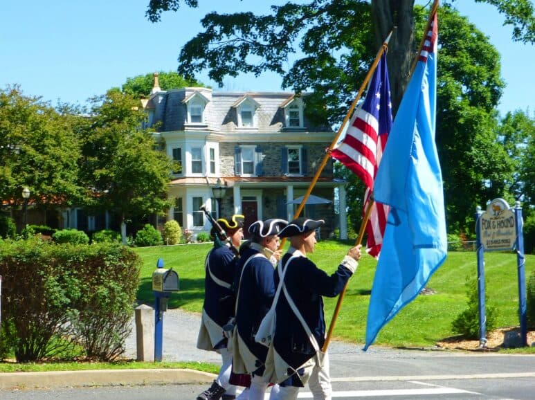 Three men in historical military uniforms march with flags in front of a large house.