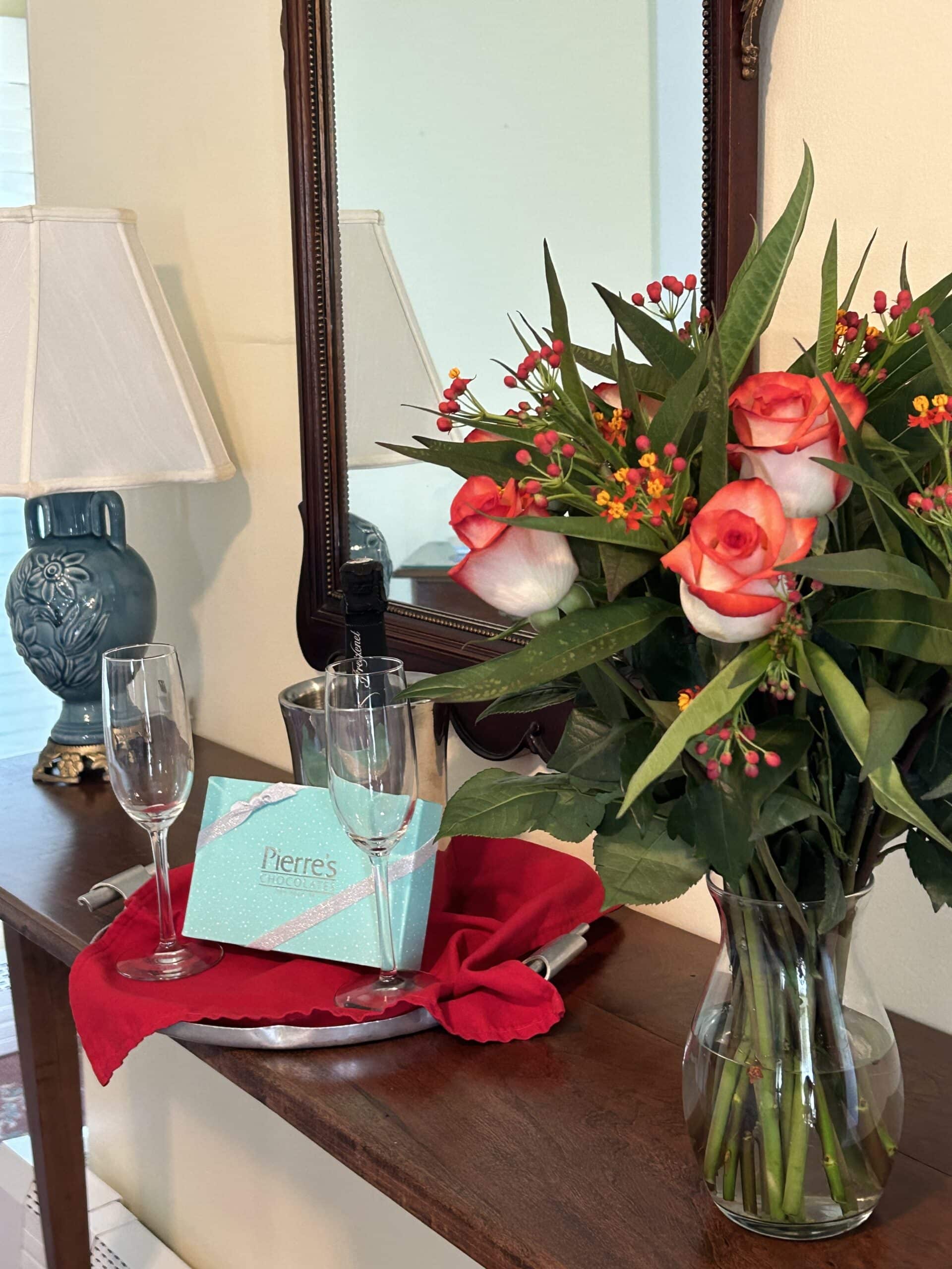 A decorative table with a lamp, a mirror, a bouquet of roses, champagne flutes, and a gift box.