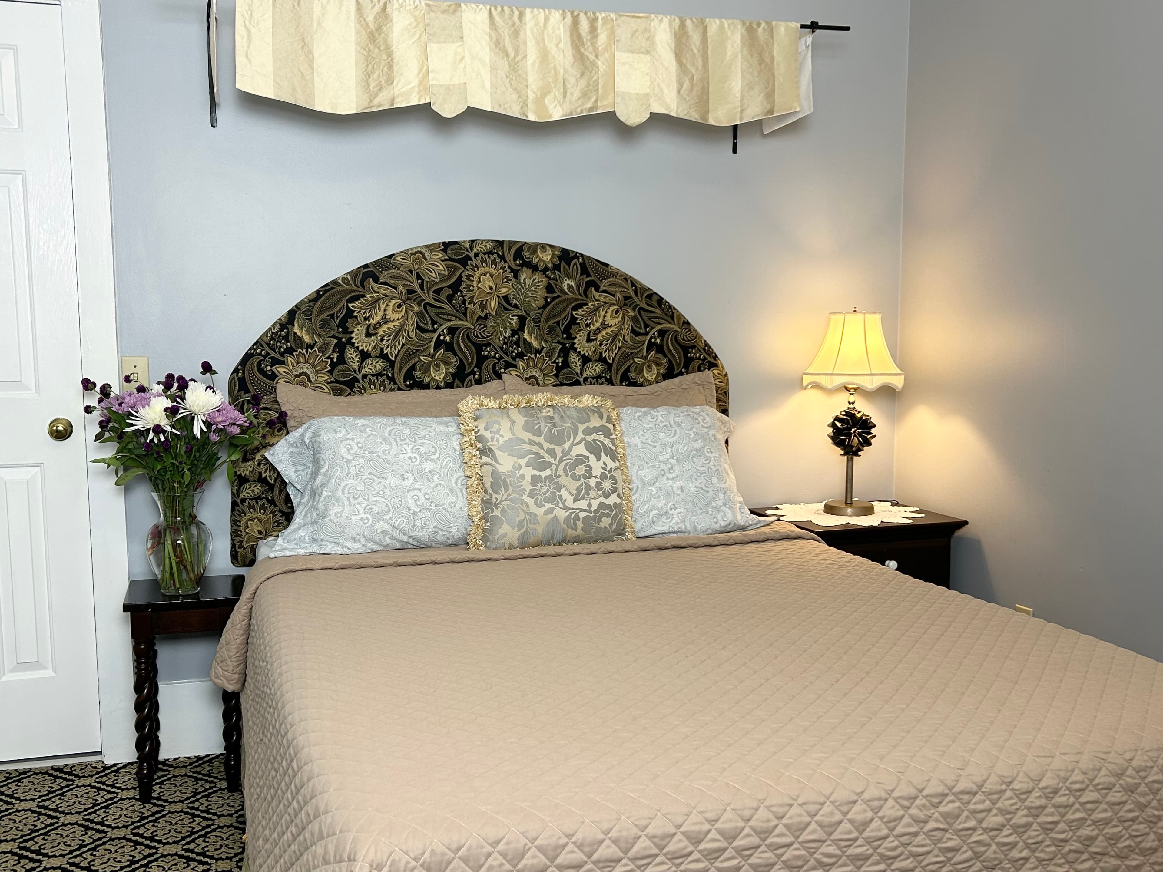A cozy bedroom featuring a neatly made bed, decorative pillows, a bedside lamp, and a vase of flowers on a nightstand.