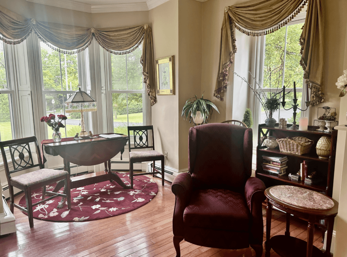 A cozy living room with large windows, a round table, two chairs, a burgundy armchair, and decorative plants.