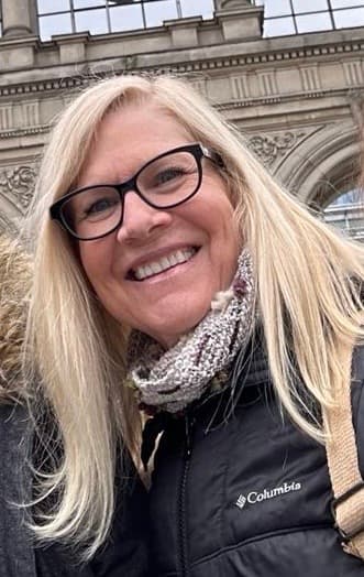 A smiling woman with long blonde hair, wearing glasses and a black jacket, stands in front of a historic building.