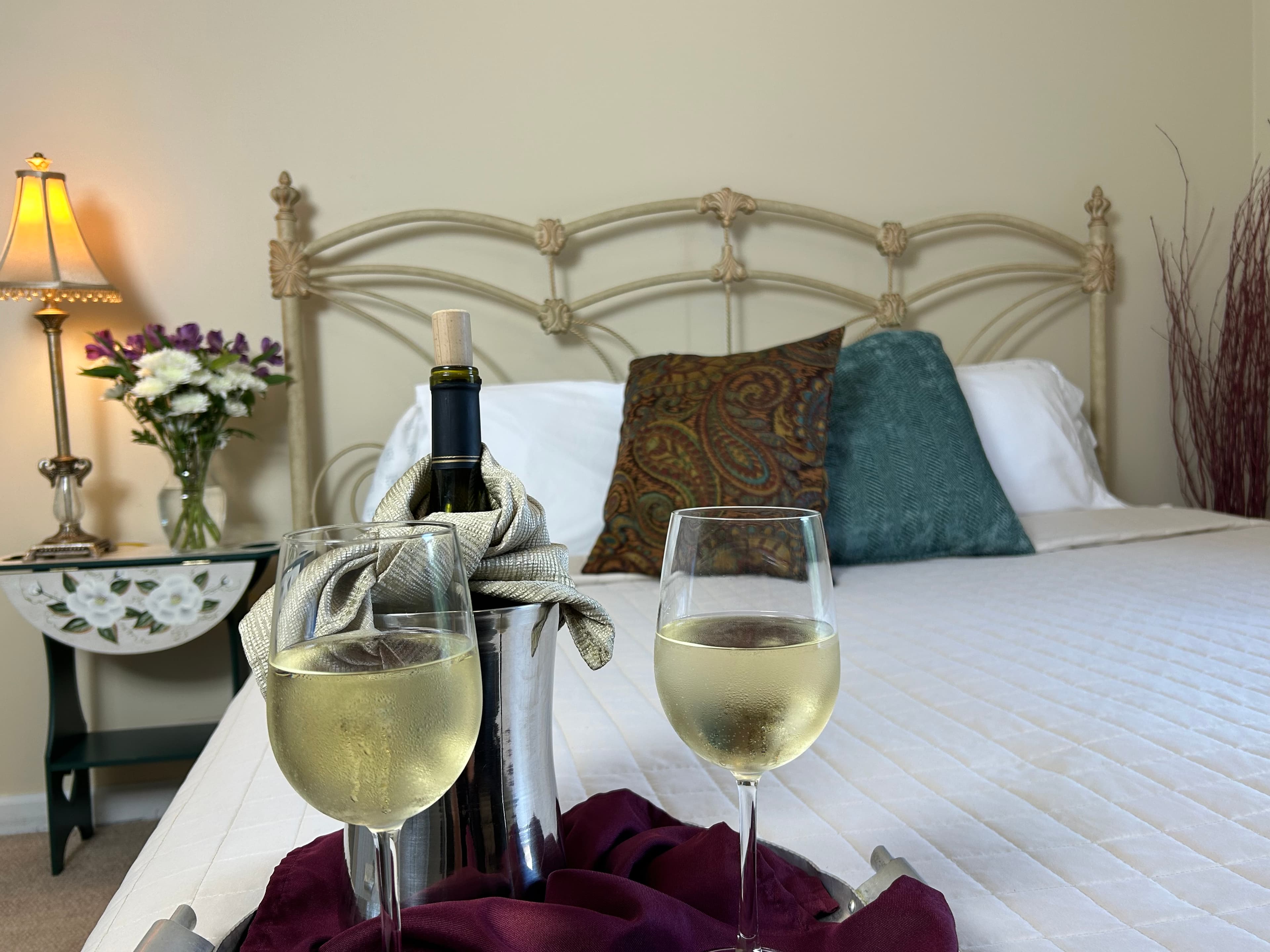 View of king bed, side table & lamp with wine & wine glasses on bed
