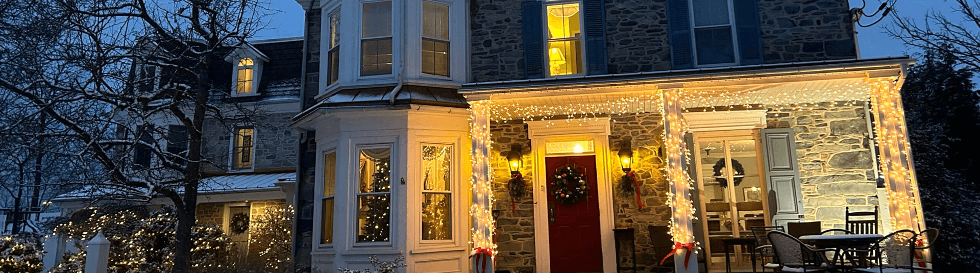 A beautifully lit stone house decorated for the holidays, surrounded by snow-covered branches.
