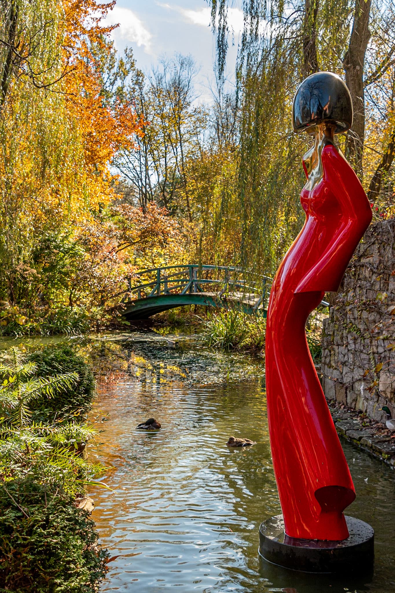 A towering red sculpture of a woman stands beside a serene pond surrounded by autumn foliage and a small bridge.